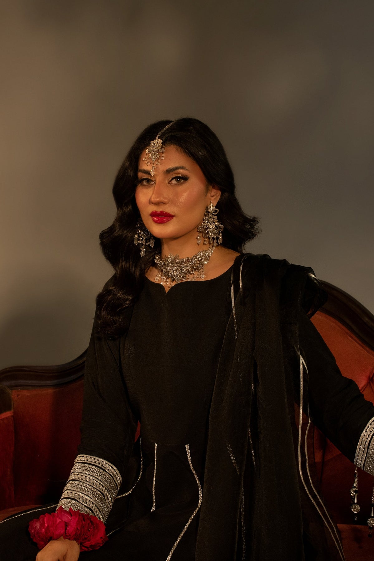 Woman in traditional attire with jewelry sitting on a red couch against a dark background
