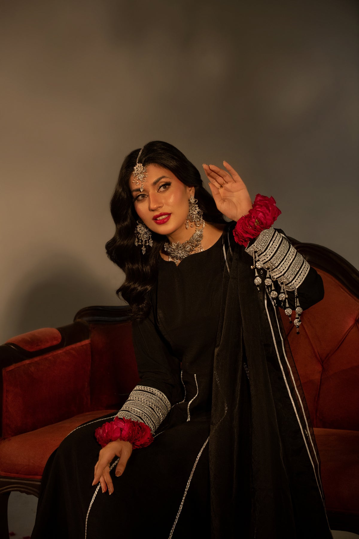 Woman in traditional attire with jewelry sitting on a red couch against a gray background