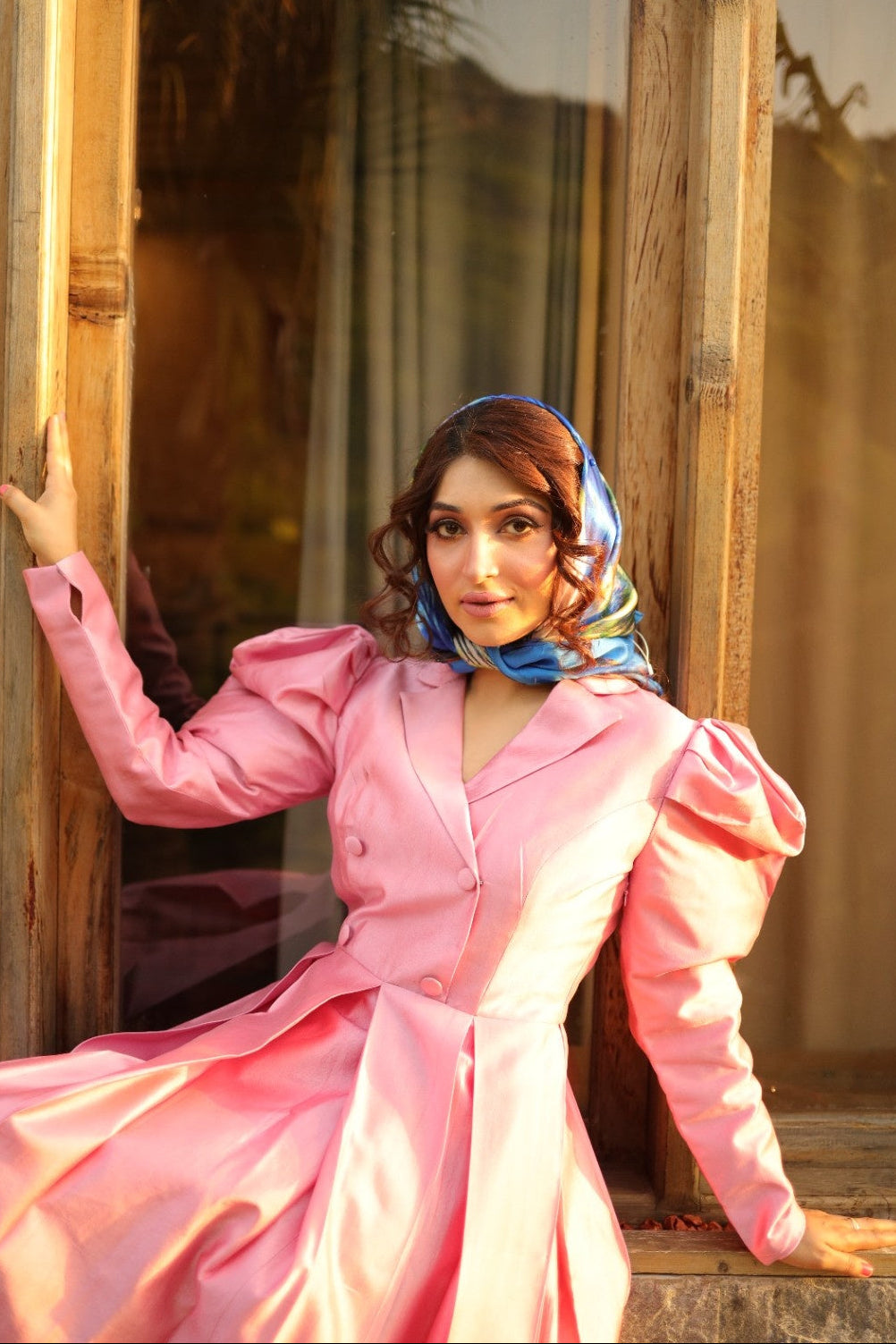 Woman in a pink dress with a blue headscarf standing in front of a wooden door.