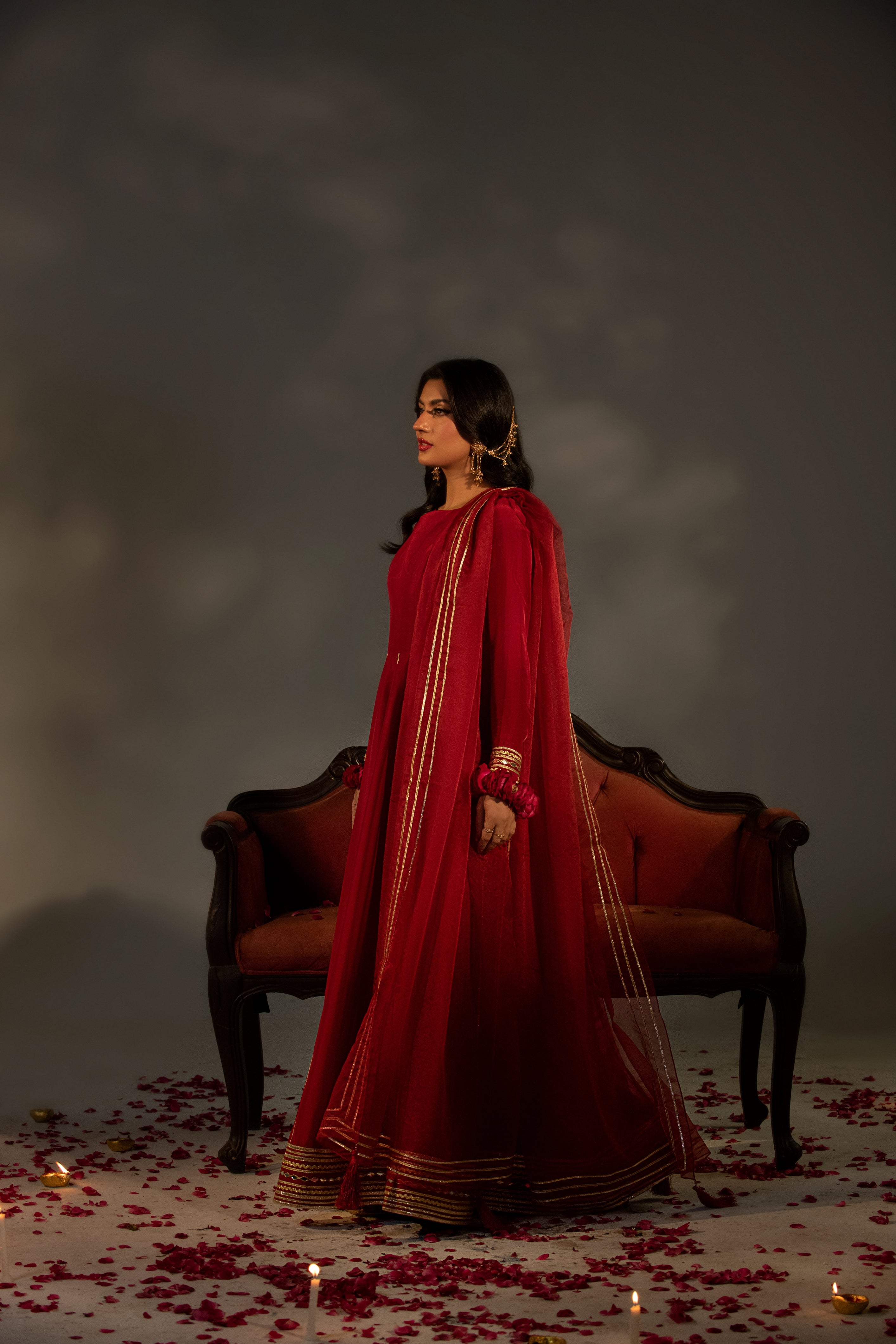 Woman in a red traditional outfit standing in front of a dark background with candles and rose petals on the floor.