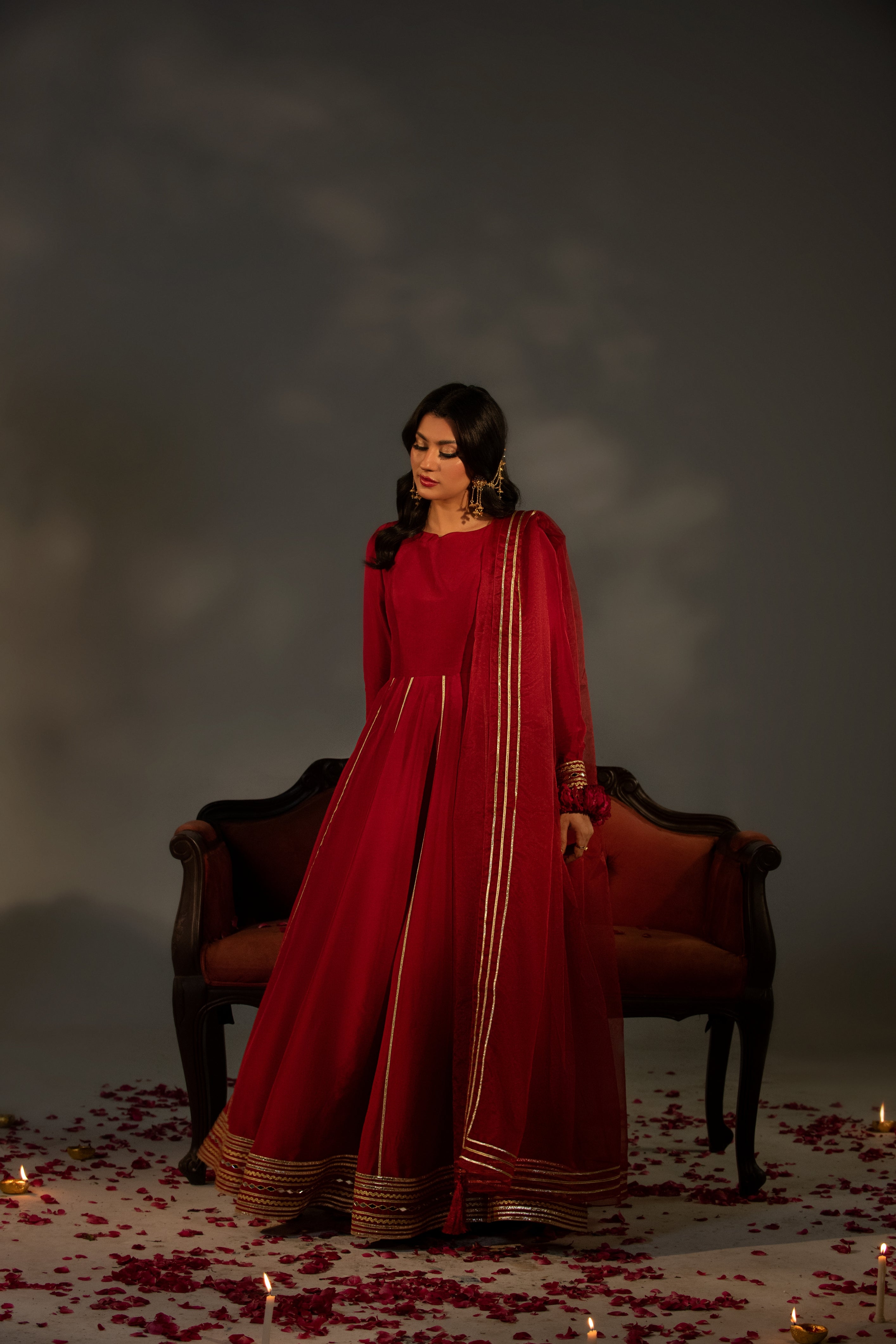 Woman in a red traditional outfit standing in front of a dark background with candles and rose petals.