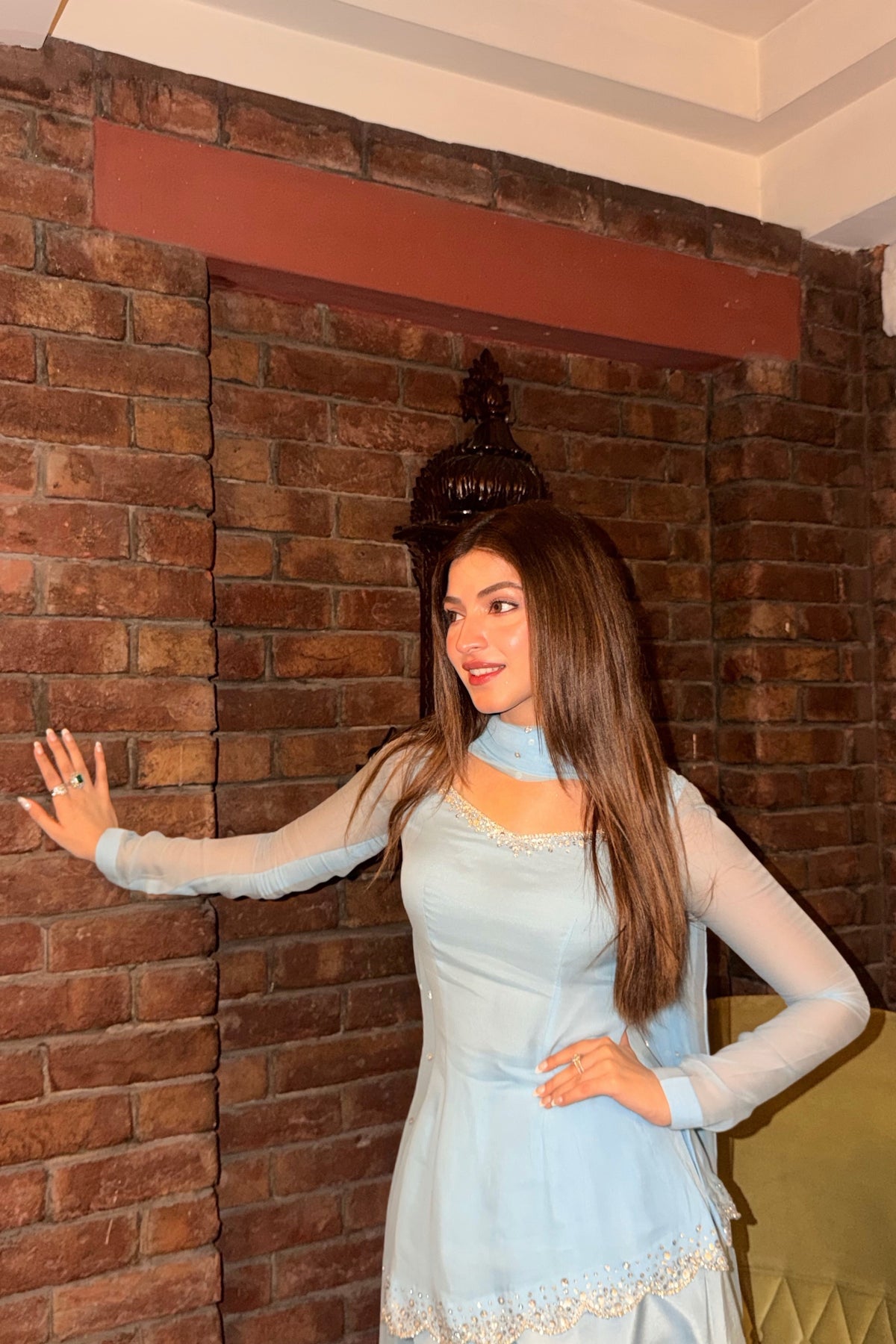 A woman posing in front of a brick wall, wearing a light blue traditional dress with sheer long sleeves and a high neck.