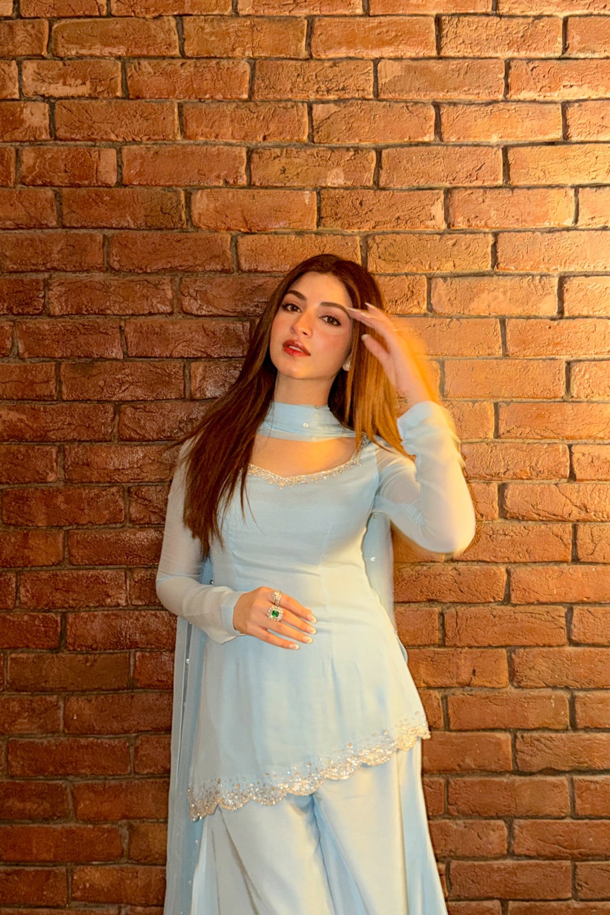 A woman posing in a light blue traditional outfit with a matching dupatta, standing against a warm-lit brick wall.