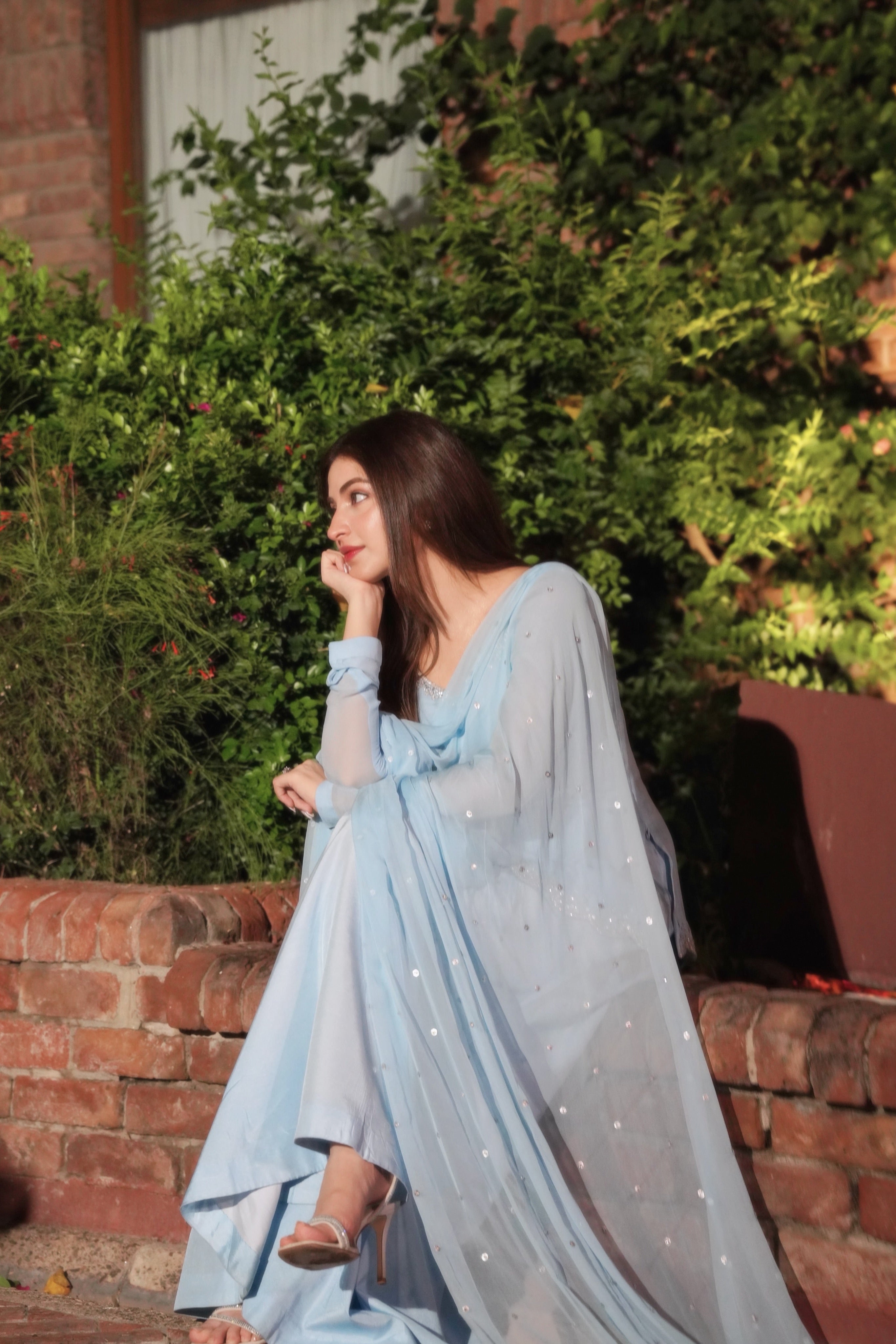 A woman wearing a light blue embroidered traditional outfit with a sheer draped shawl sits on a brick ledge in front of green foliage and a brick wall.