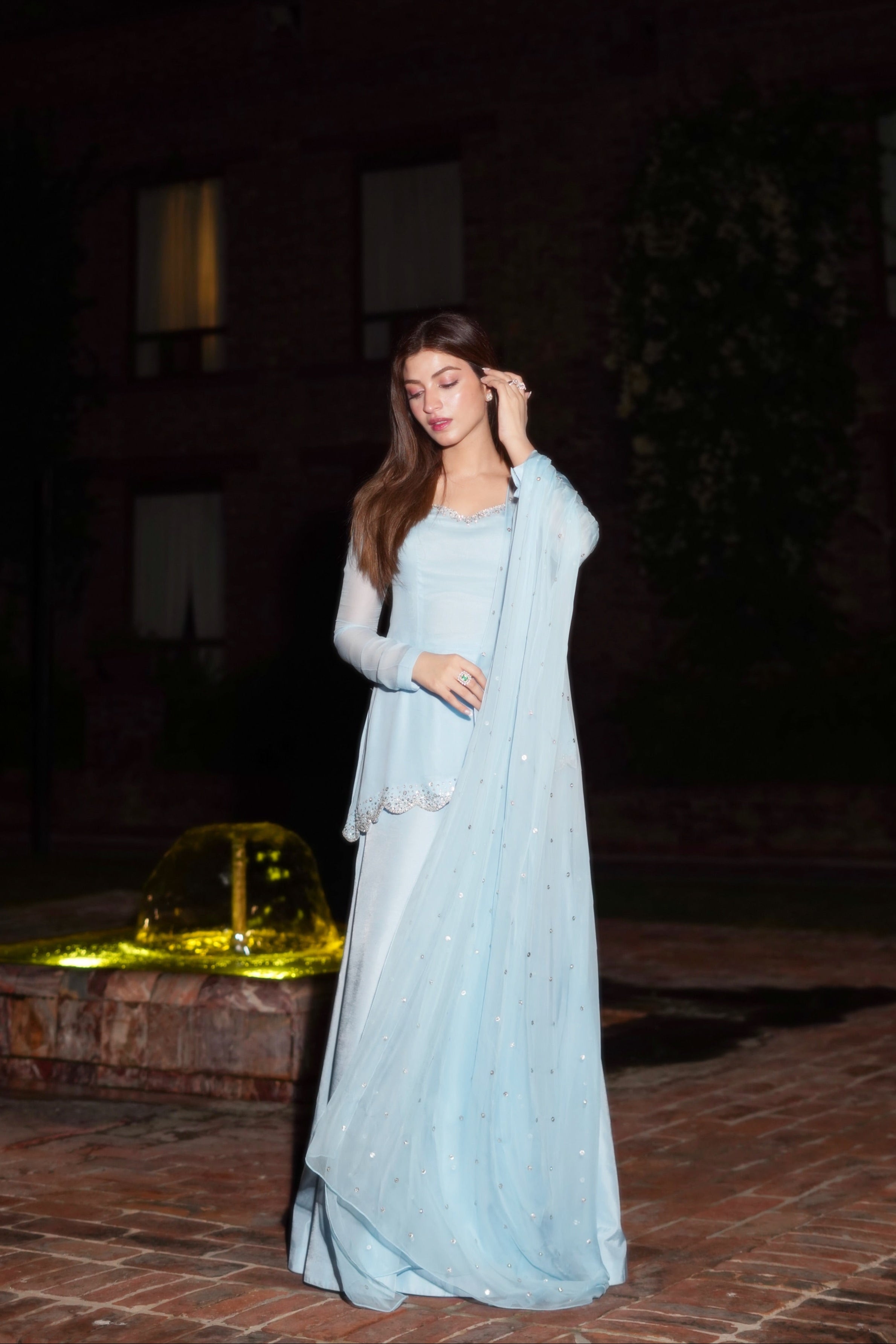 A woman wearing a light blue traditional gown with a matching dupatta stands outdoors at night on a brick patio in front of a building with lit windows and a fountain in the background.