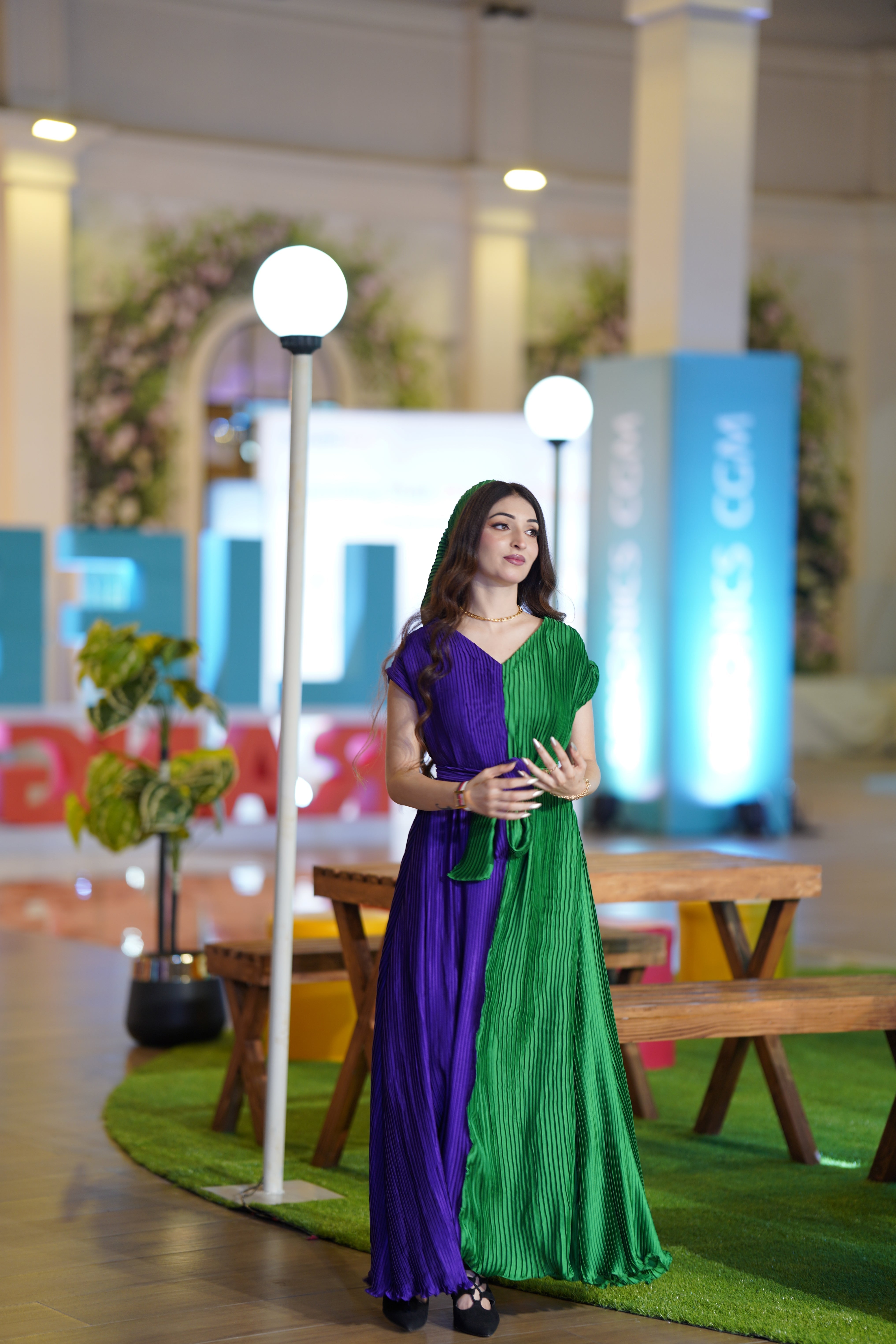 A woman wearing a long, pleated two-tone dress in purple and green stands on a green carpeted display area with wooden benches, lamp posts, and blue signage in the background.
