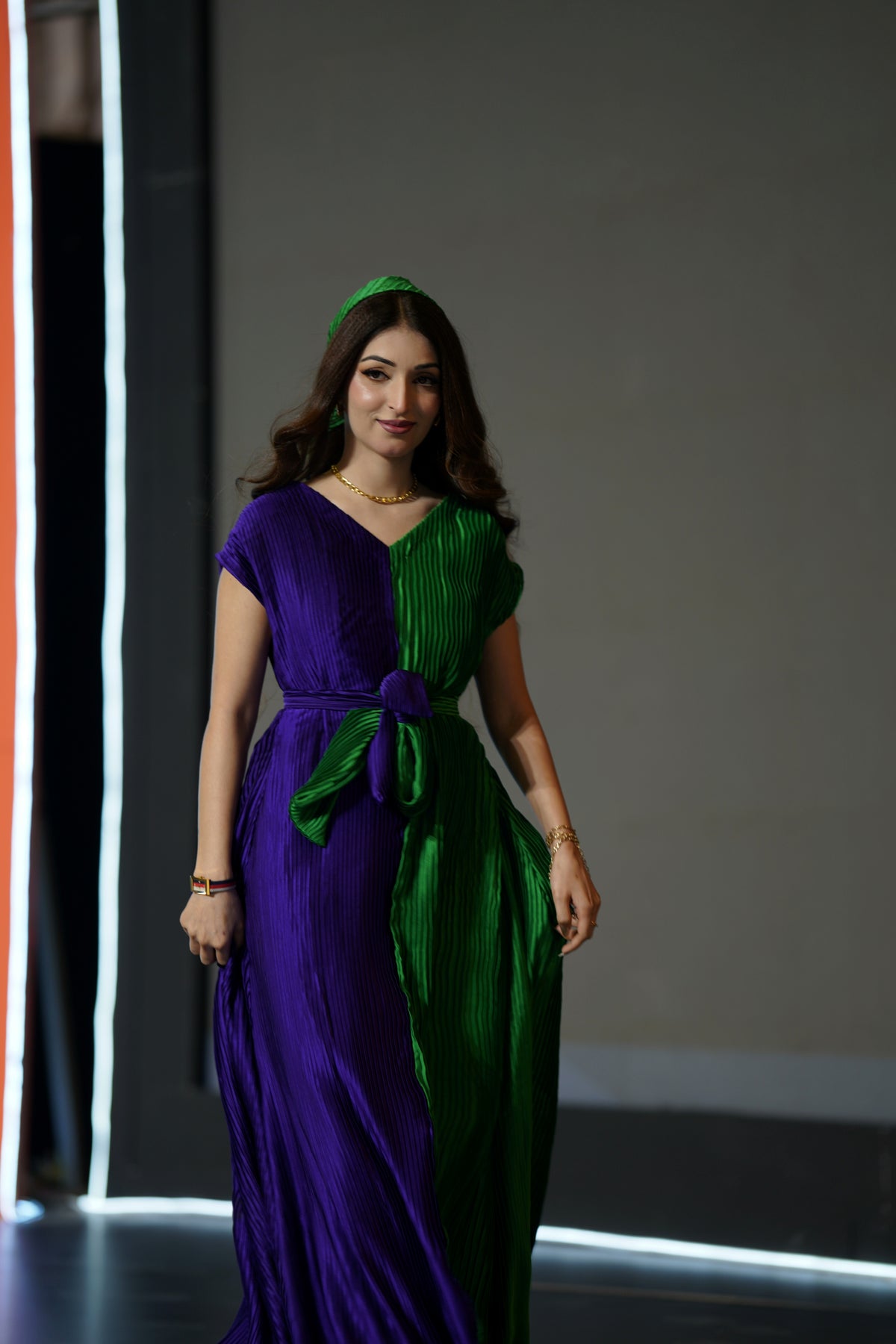 A woman modeling a floor-length pleated dress with a split purple and green color-block design, featuring a matching green headband, set against a dark gray runway background with illuminated vertical lines.
