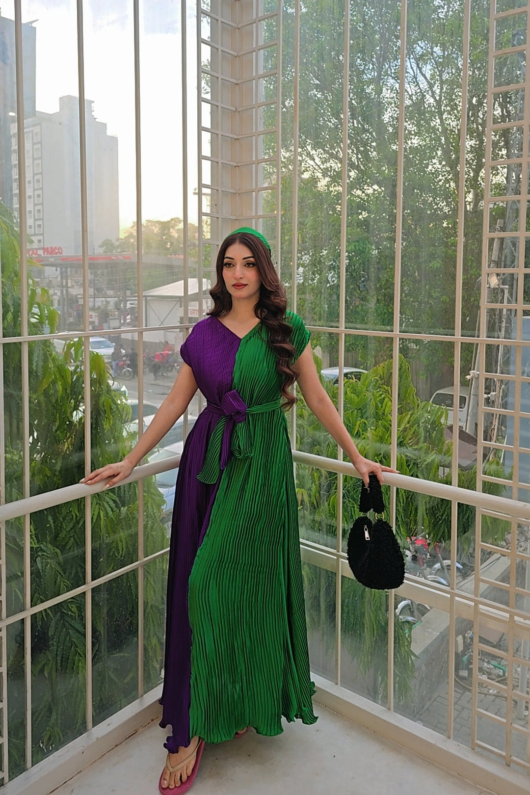 A woman stands in a glass-walled sunroom wearing a long green and purple pleated dress with a matching headband, holding a black handbag.
