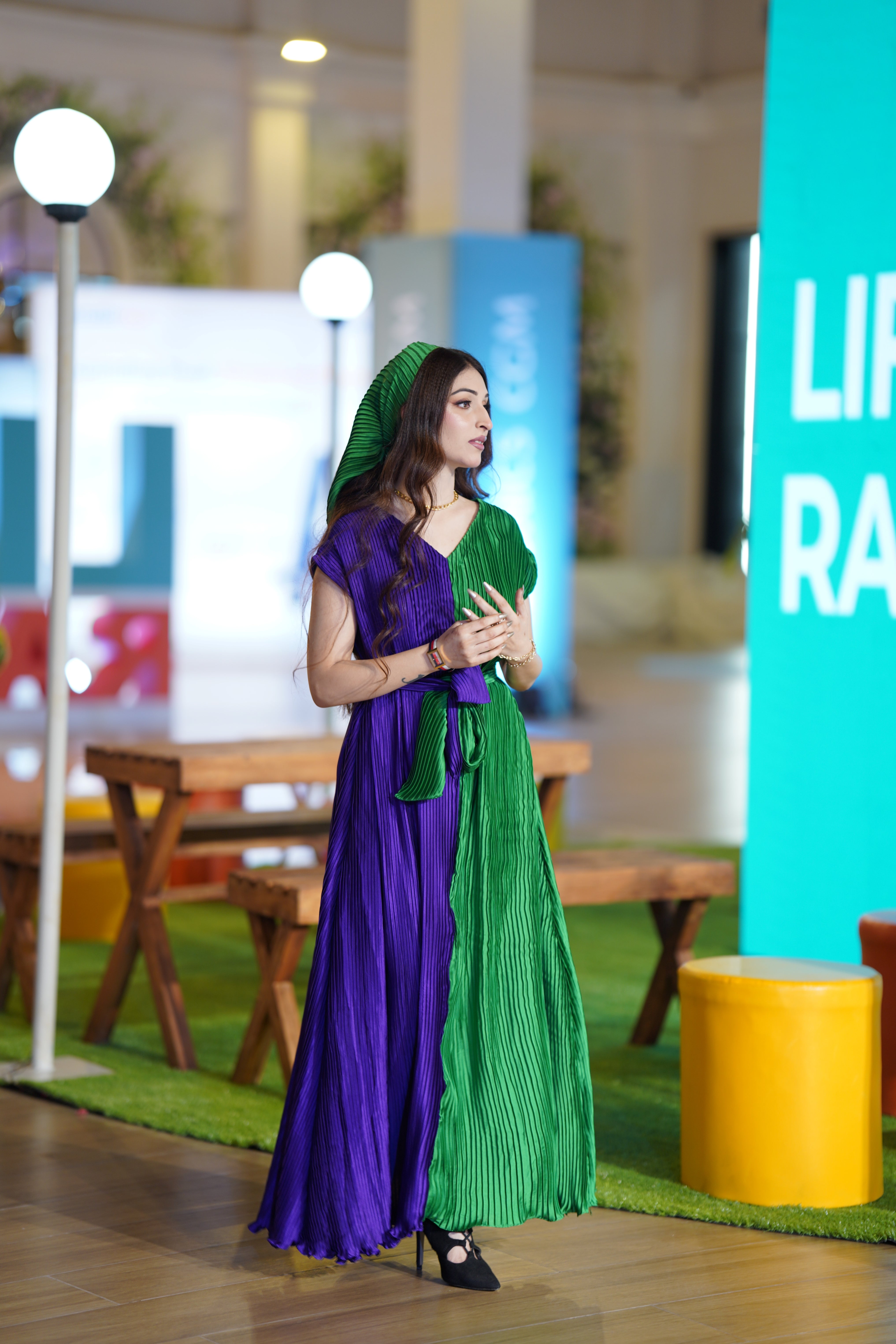 A woman stands indoors wearing a long pleated dress split vertically into purple and green sections, with a matching green head covering. The setting appears to be a styled event or exhibition space with tables, lamps, a bright blue panel, and a yellow stool in the background.
