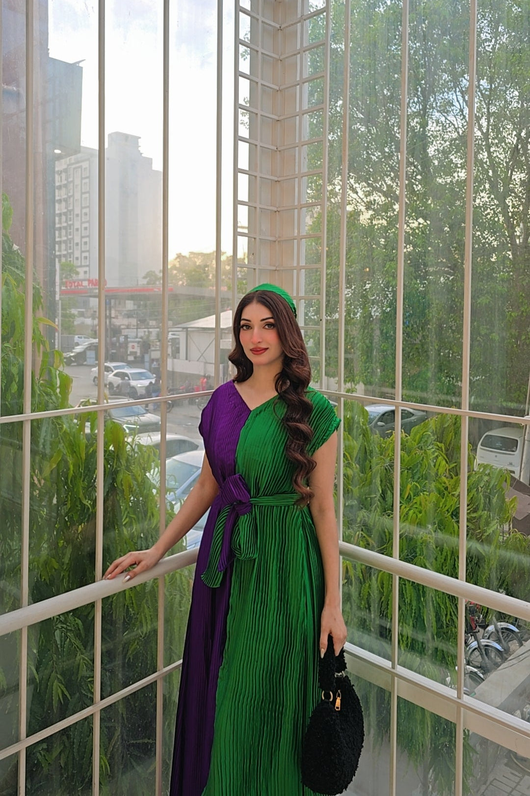 A woman wearing a long pleated dress with a purple and green color-blocked design stands in a glass-walled room with an outdoor city view. She is holding a black handbag.