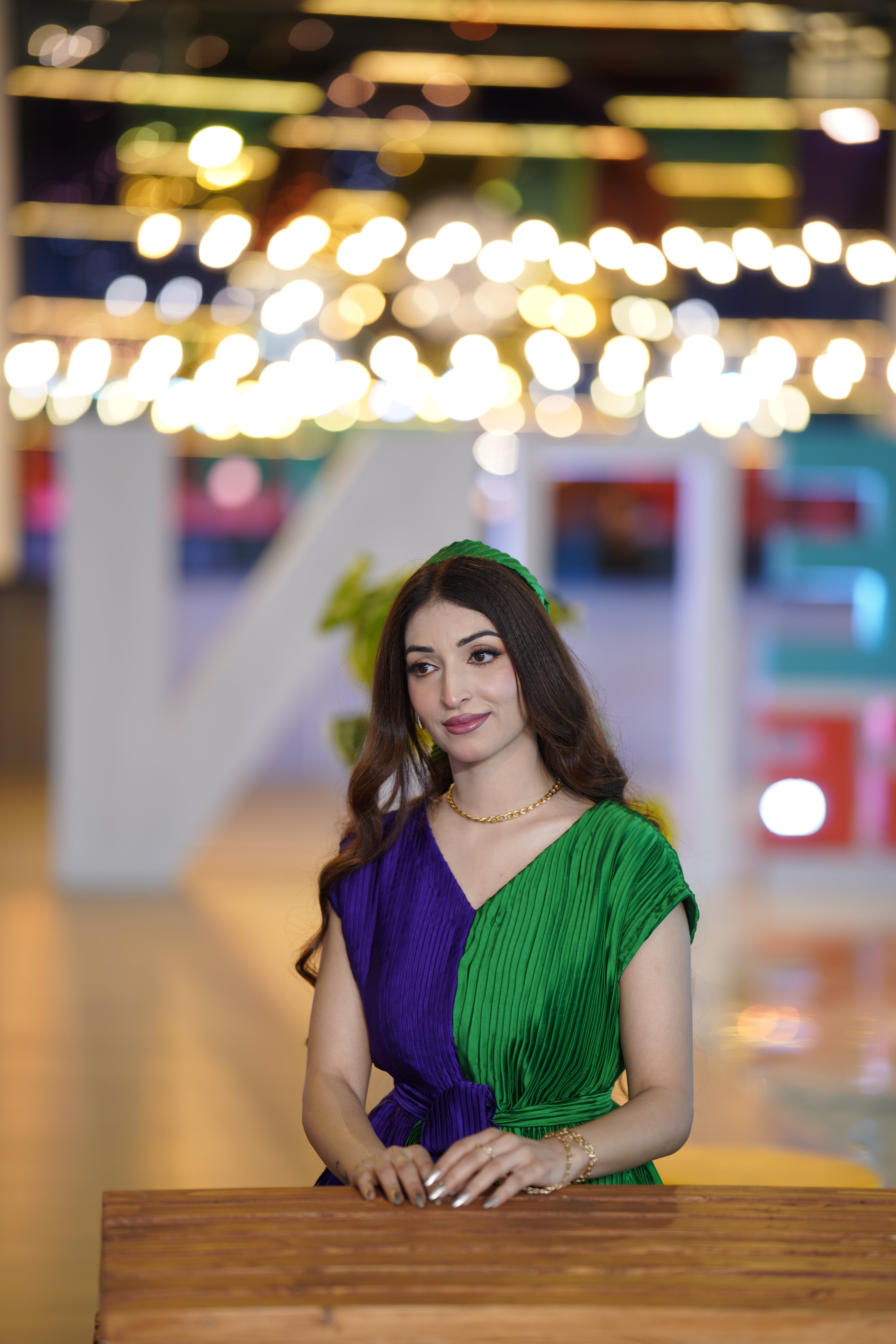 A woman wearing a pleated two-tone dress in purple and green, sitting behind a wooden table in a brightly lit indoor setting with blurred lights in the background.