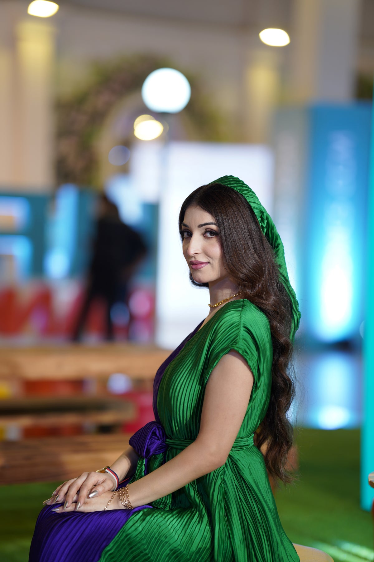 A woman wearing a vibrant green pleated dress with a matching green headscarf and a purple sash, seated in a brightly lit, blurred indoor event space.