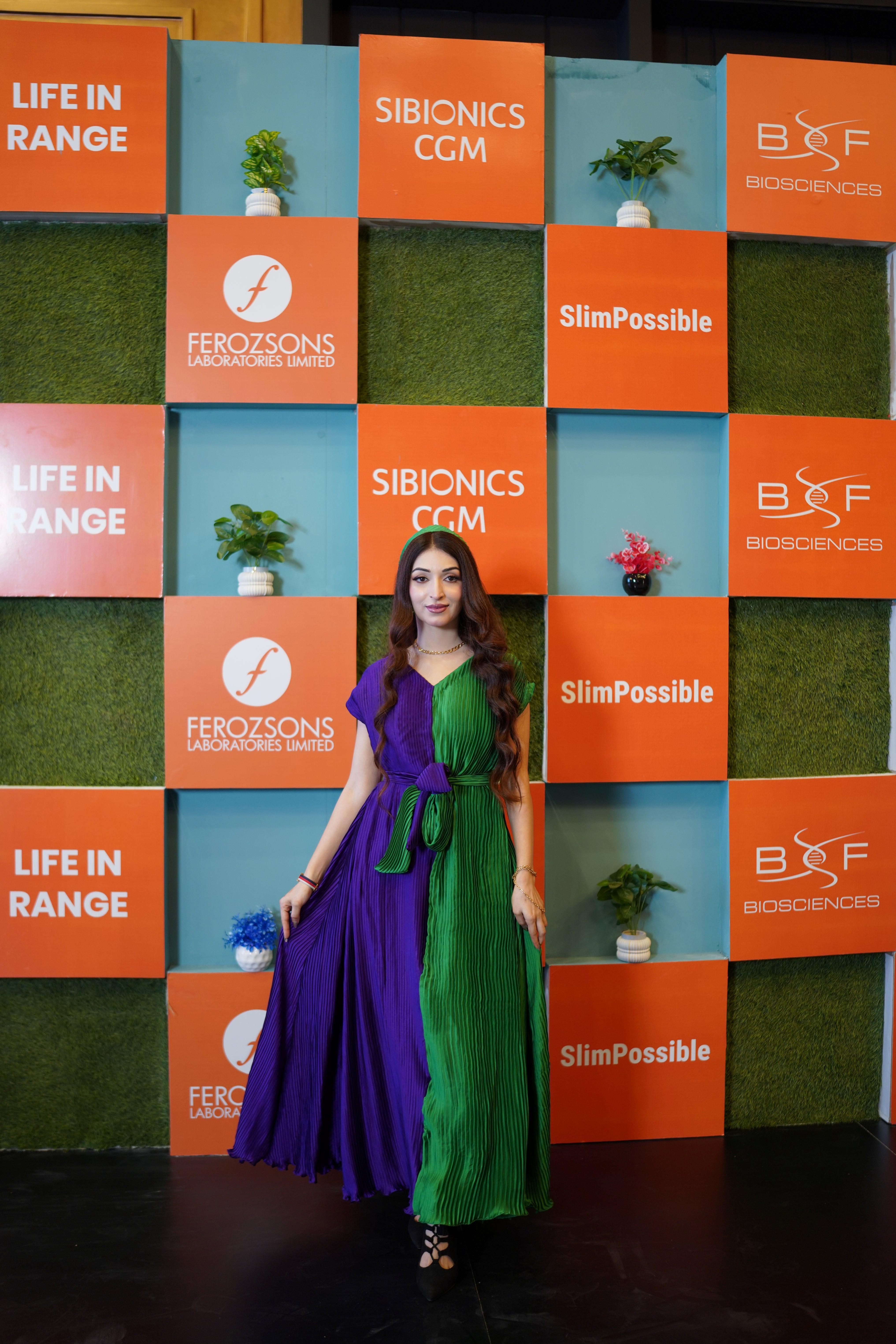A woman stands in front of a branded backdrop wall wearing a long pleated dress split vertically into purple on the left and green on the right. The wall features orange and blue panels with logos including Sibionics CGM, Ferozsons Laboratories, and BNF BioSciences.