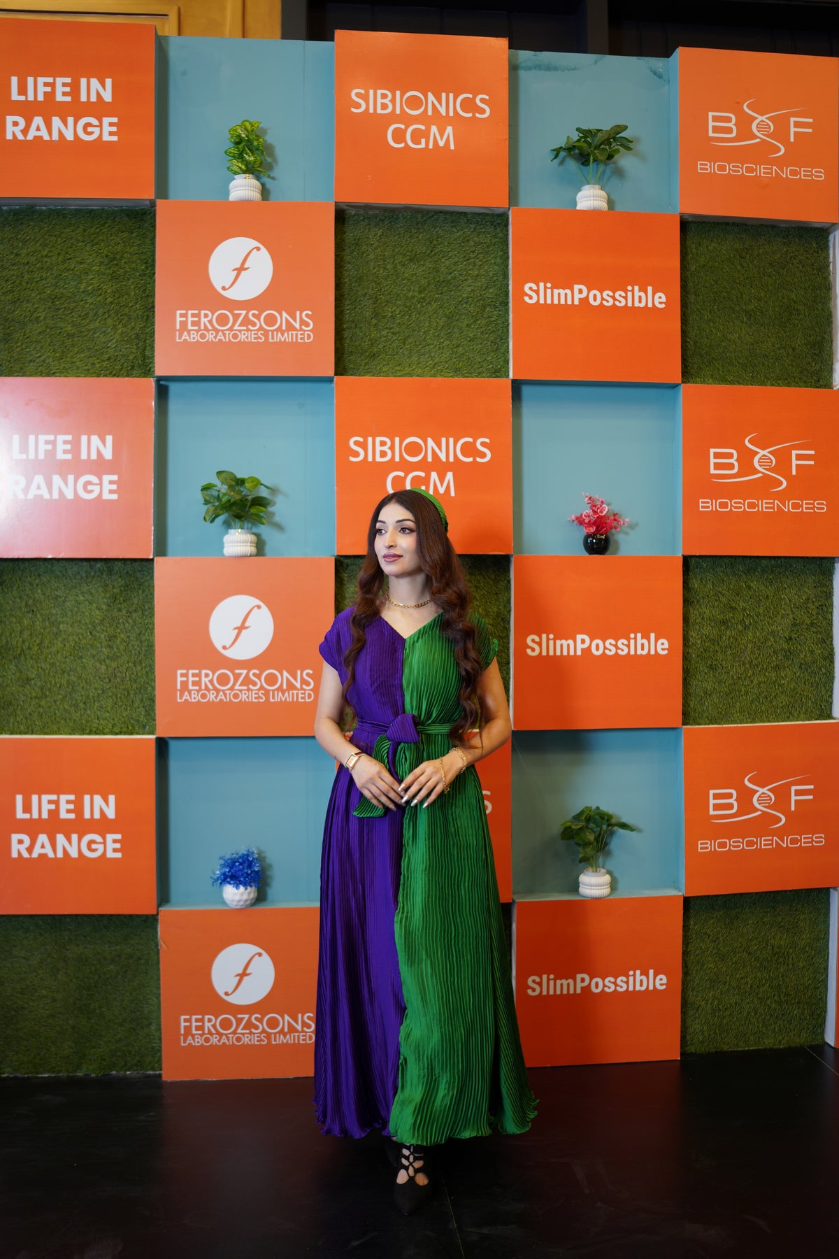 A woman wearing a pleated two-tone dress in purple and green stands in front of a branded backdrop wall featuring logos for Ferazons, Sibionics CCM, and BCF Biosciences.