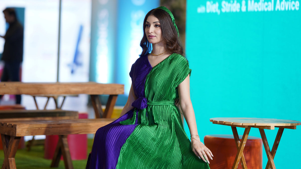 A woman wearing a floor-length pleated dress with a split green and purple color-block design sits on a green carpeted runway. A large teal backdrop on the right displays the text '#SlimPossible with Diet, Stride & Medical Advice'.