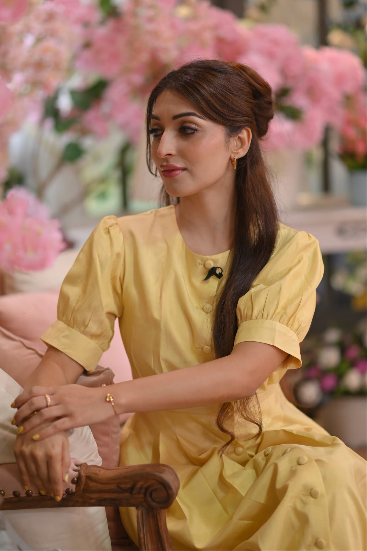 Woman in a yellow dress sitting in a decorated indoor setting with pink flowers.