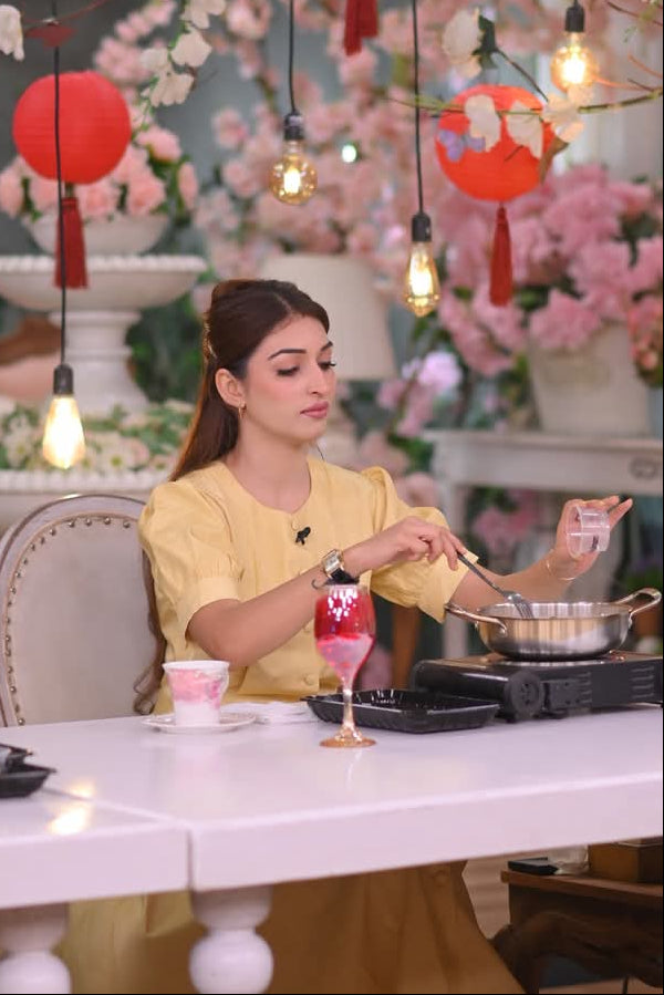 Woman in a yellow outfit preparing food at a table with decorative lights and flowers in the background