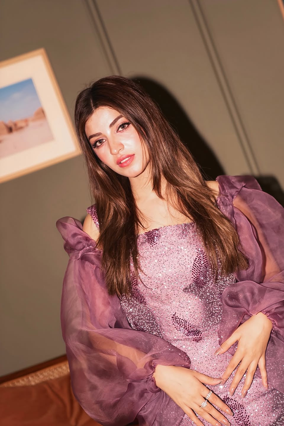 Woman in a sparkling purple dress posing indoors with framed pictures on the wall.