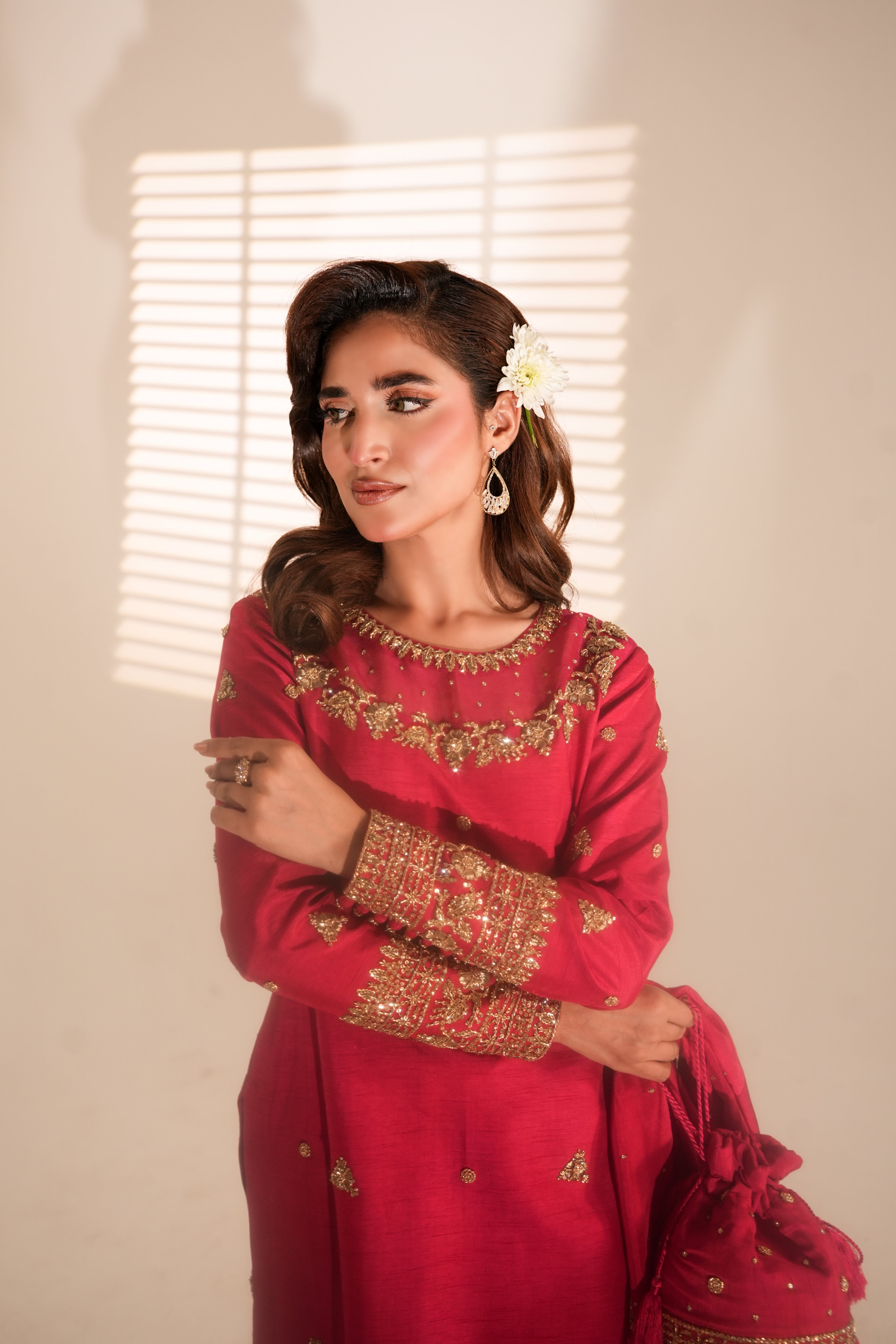 A woman models a vibrant red traditional outfit with intricate gold embroidery on the neckline and cuffs, holding a matching red embroidered clutch bag against a warm beige wall with window blinds casting soft shadows.