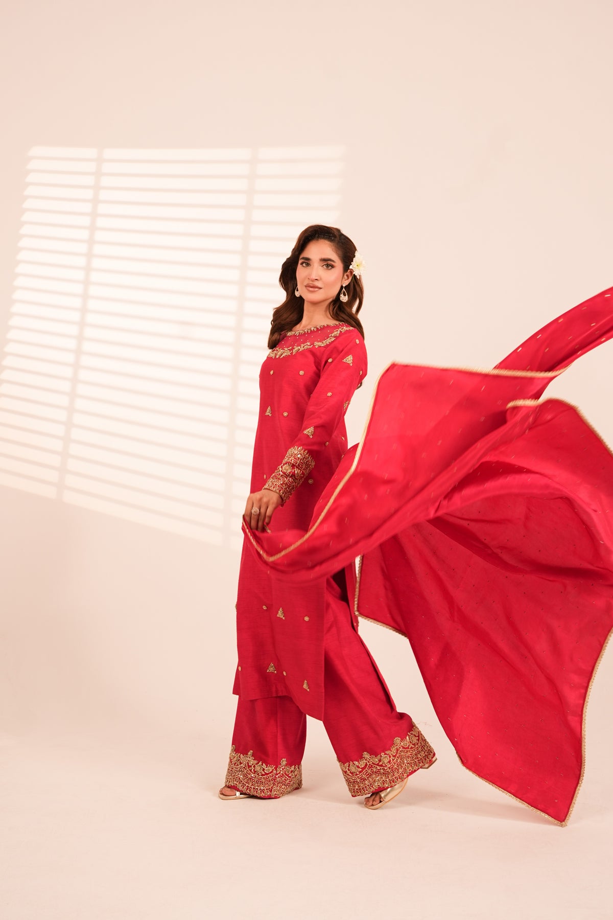 A woman modeling a bright red traditional outfit with gold embroidery and a matching flowing dupatta, posed against a warm off-white studio background with window blinds casting soft shadows.