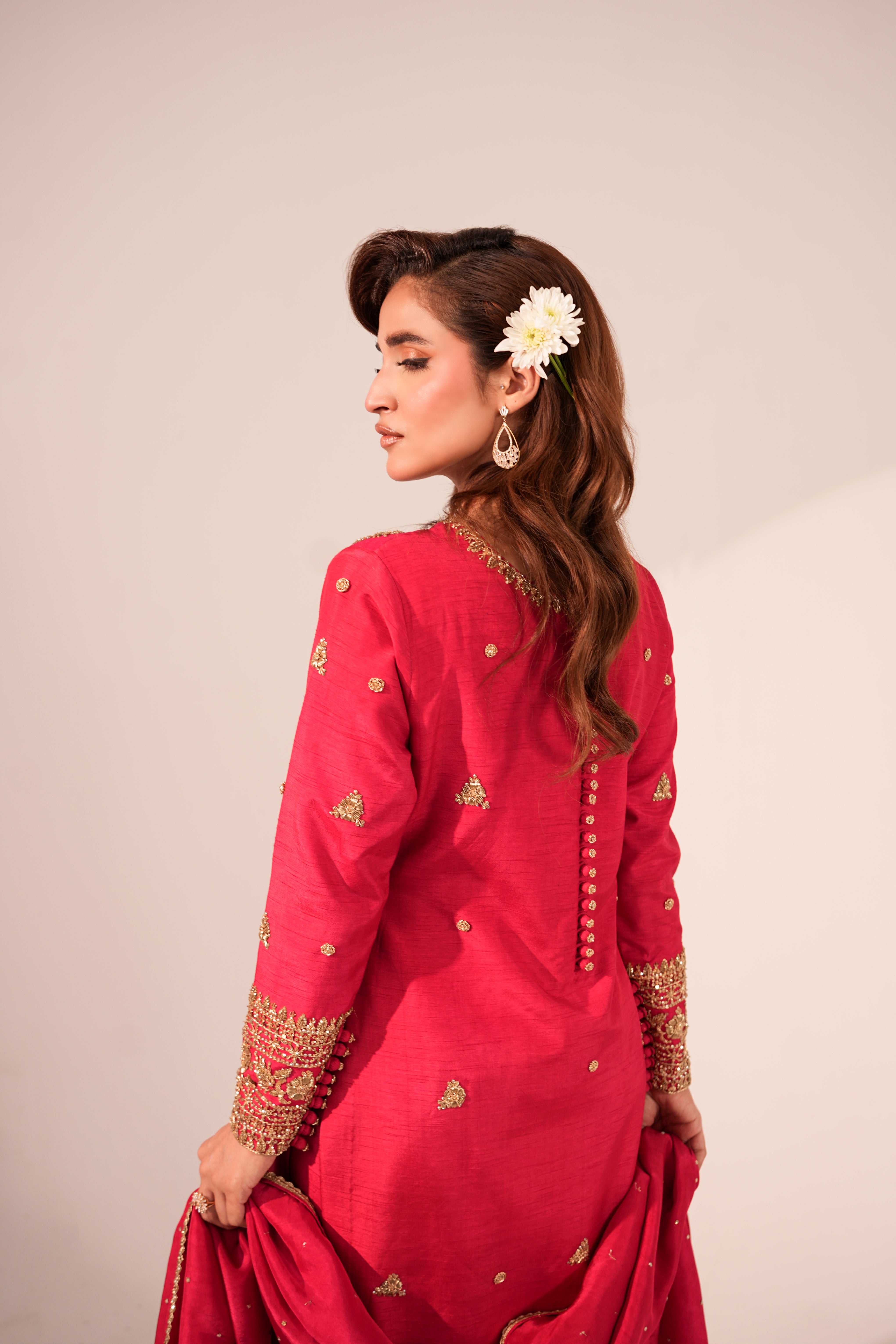 A woman wearing a bright pink traditional outfit with gold embroidery, shown from the back against a plain beige studio background.