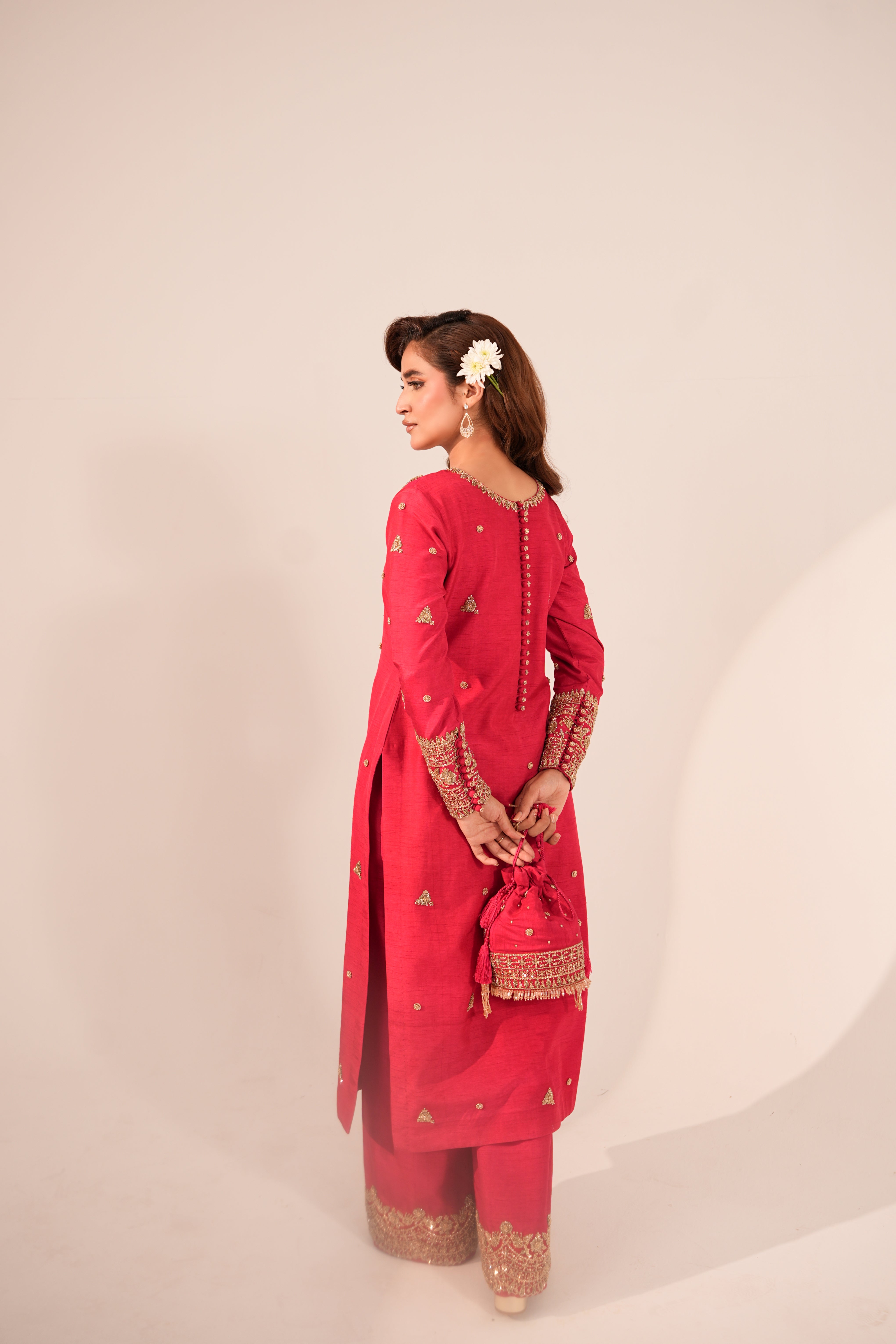 A woman modeling a bright pink traditional South Asian outfit with gold embroidery, standing in profile against a plain light beige studio background.