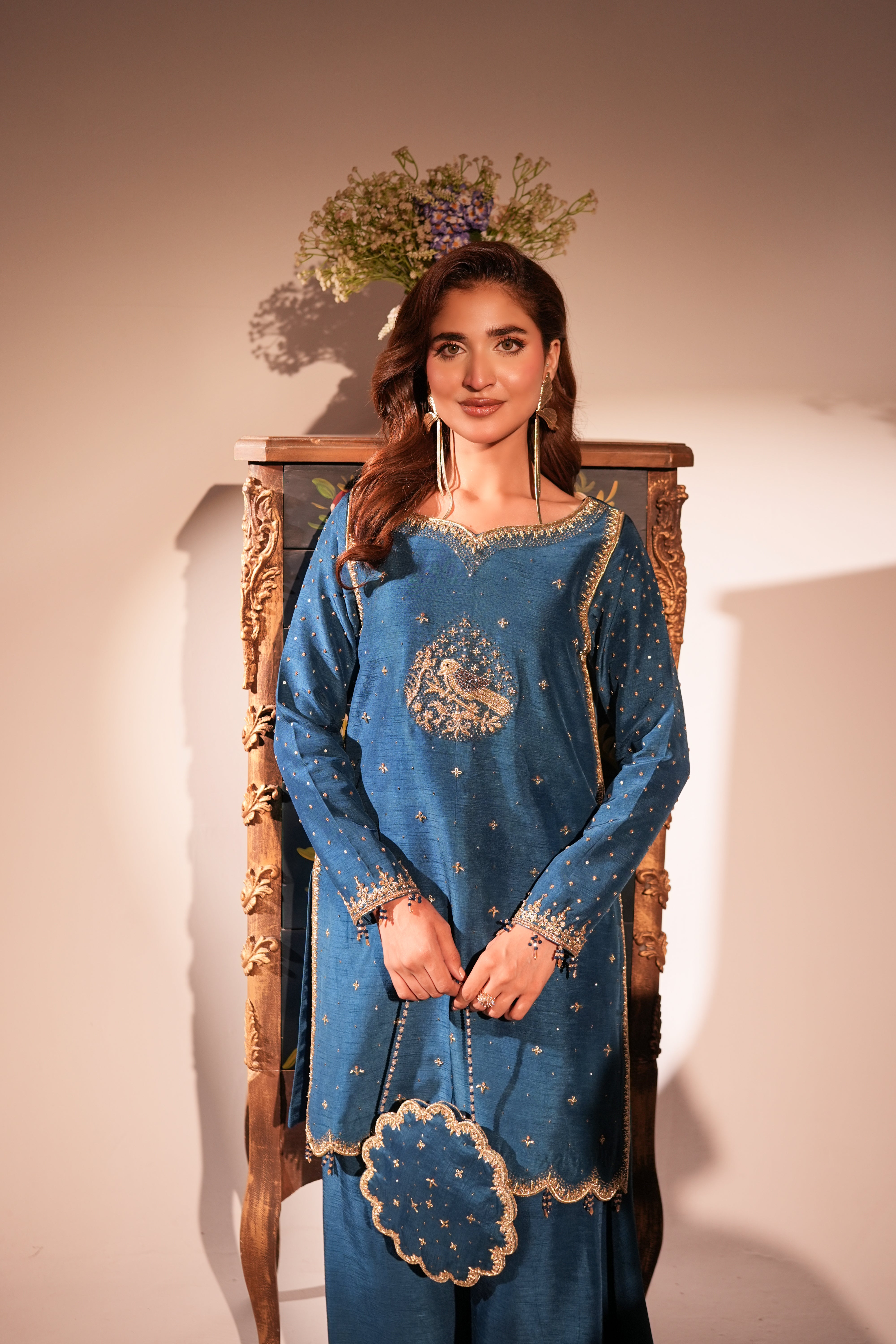 A woman models a blue traditional embroidered outfit with gold detailing, standing in front of a wooden chair against a beige wall.