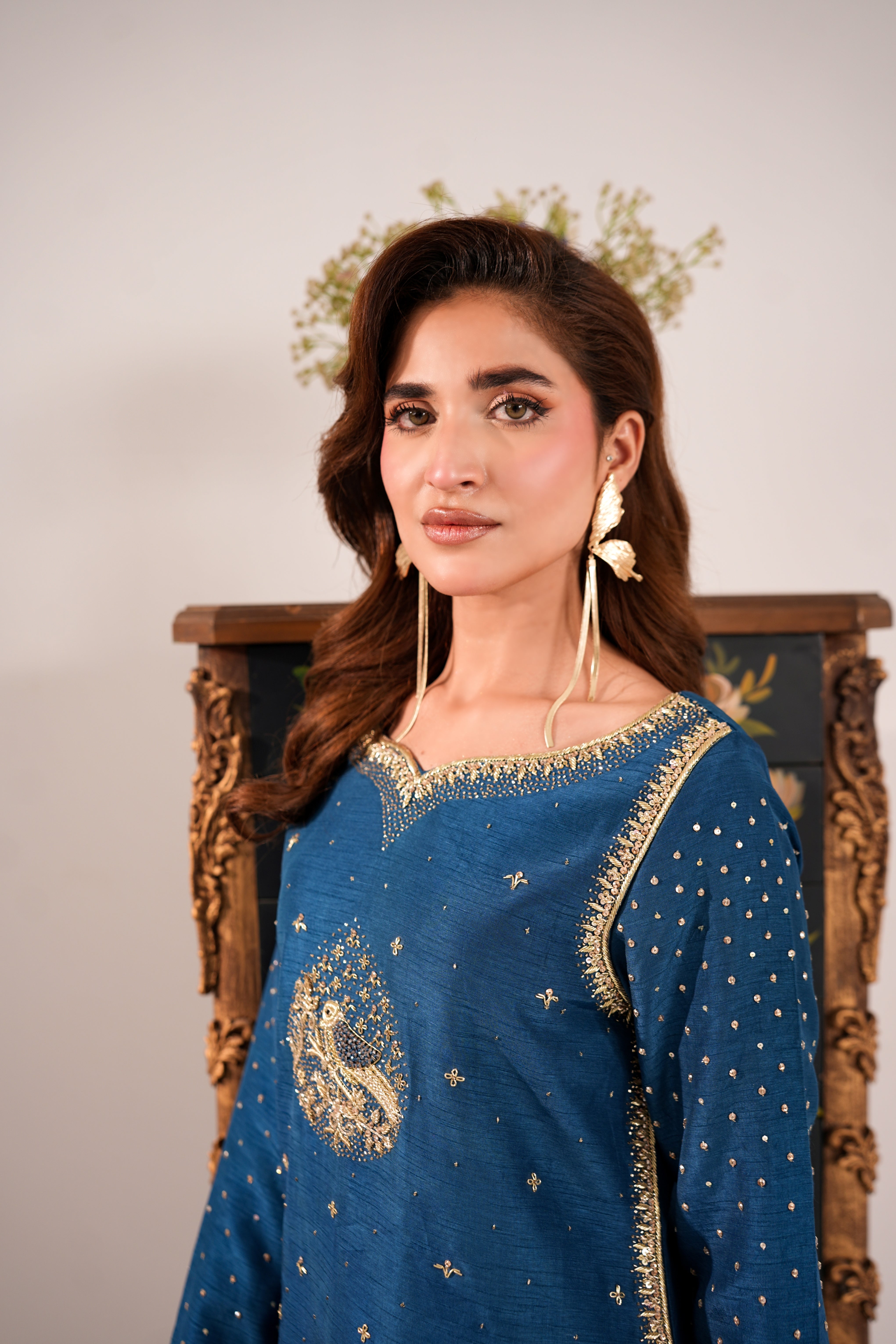 A woman wearing a blue embroidered traditional outfit with gold detailing, seated in front of an ornate wooden chair against a light neutral background.