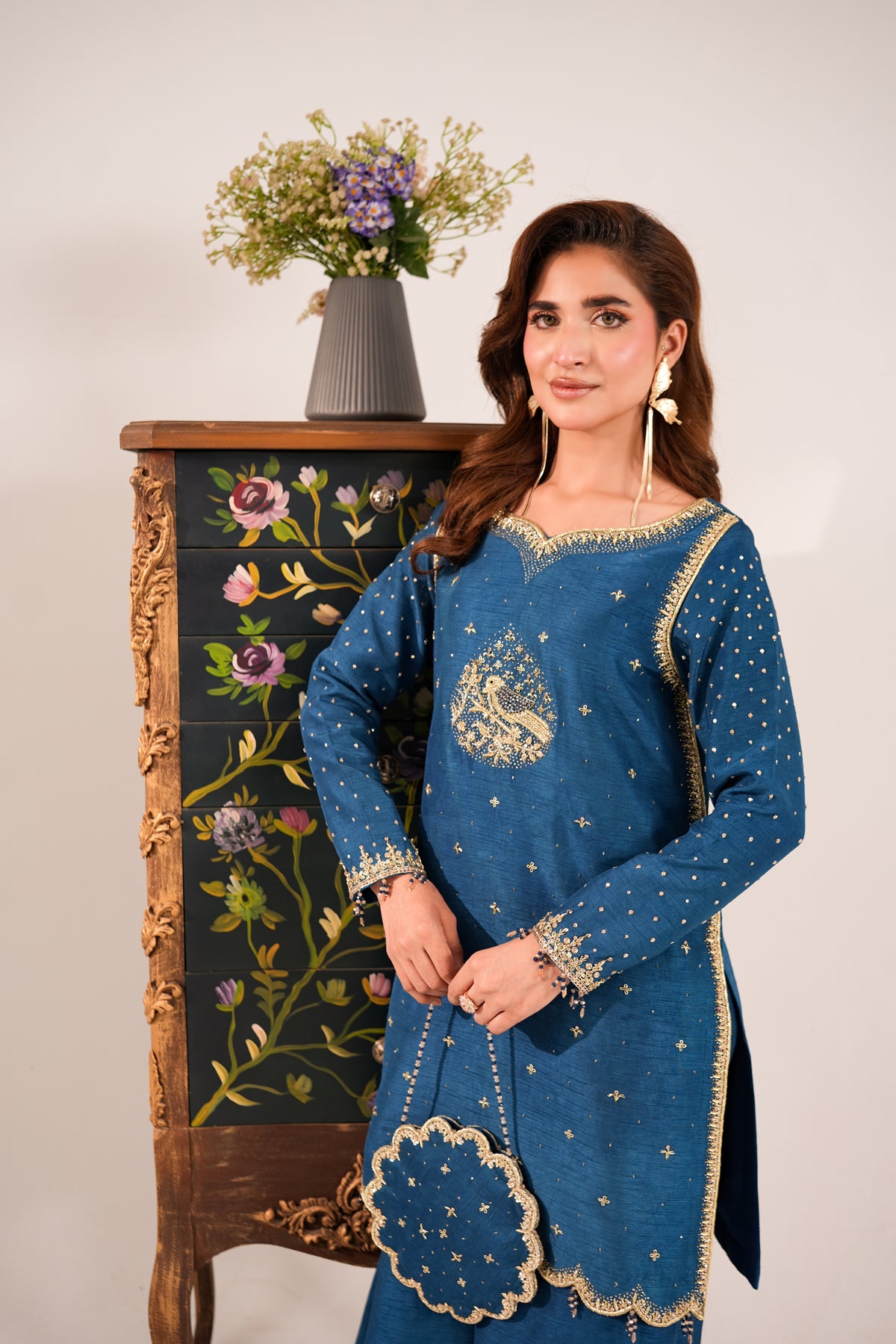 A woman models a blue traditional South Asian salwar kameez set with gold embroidery and metallic embellishments, standing in front of an ornate black cabinet with floral details.