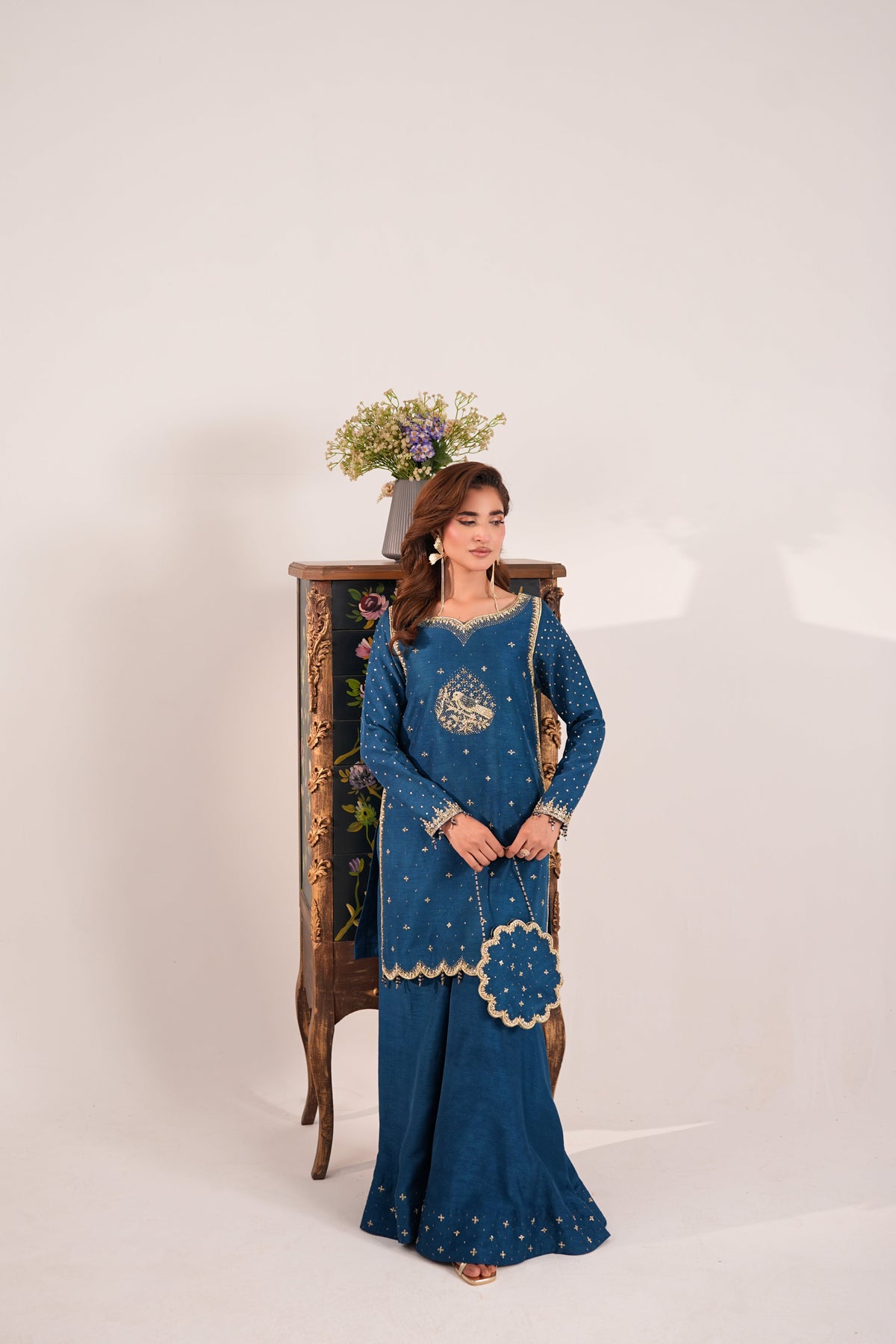 A woman models a traditional Indian salwar kameez suit in deep teal blue with gold embroidery, standing next to an ornate wooden cabinet against a plain off-white background.