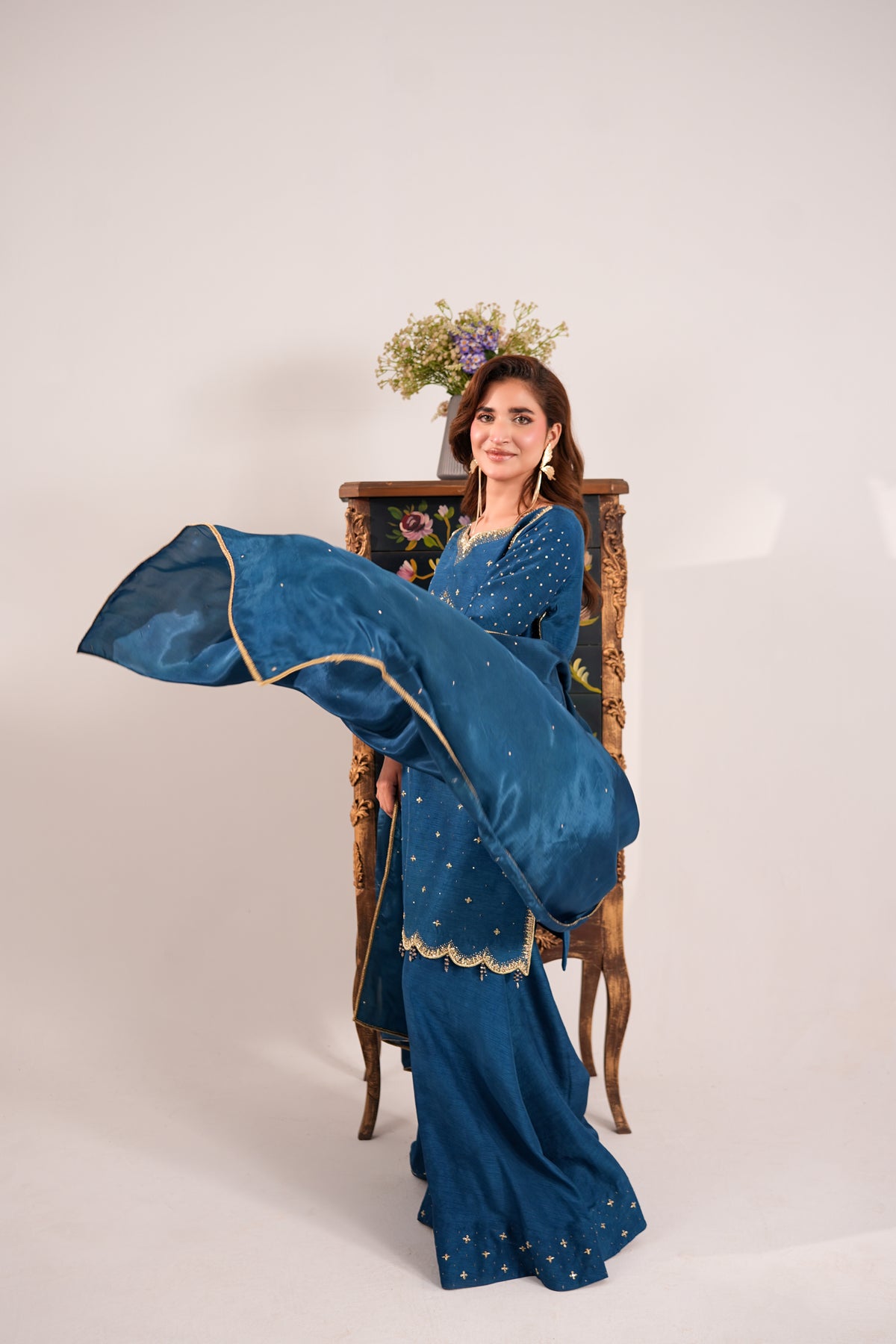 A woman models a deep teal blue traditional South Asian outfit with gold embroidery, holding a matching dupatta outstretched. She is posed in front of a vintage wooden cabinet with flowers on top, set against a plain light gray studio background.