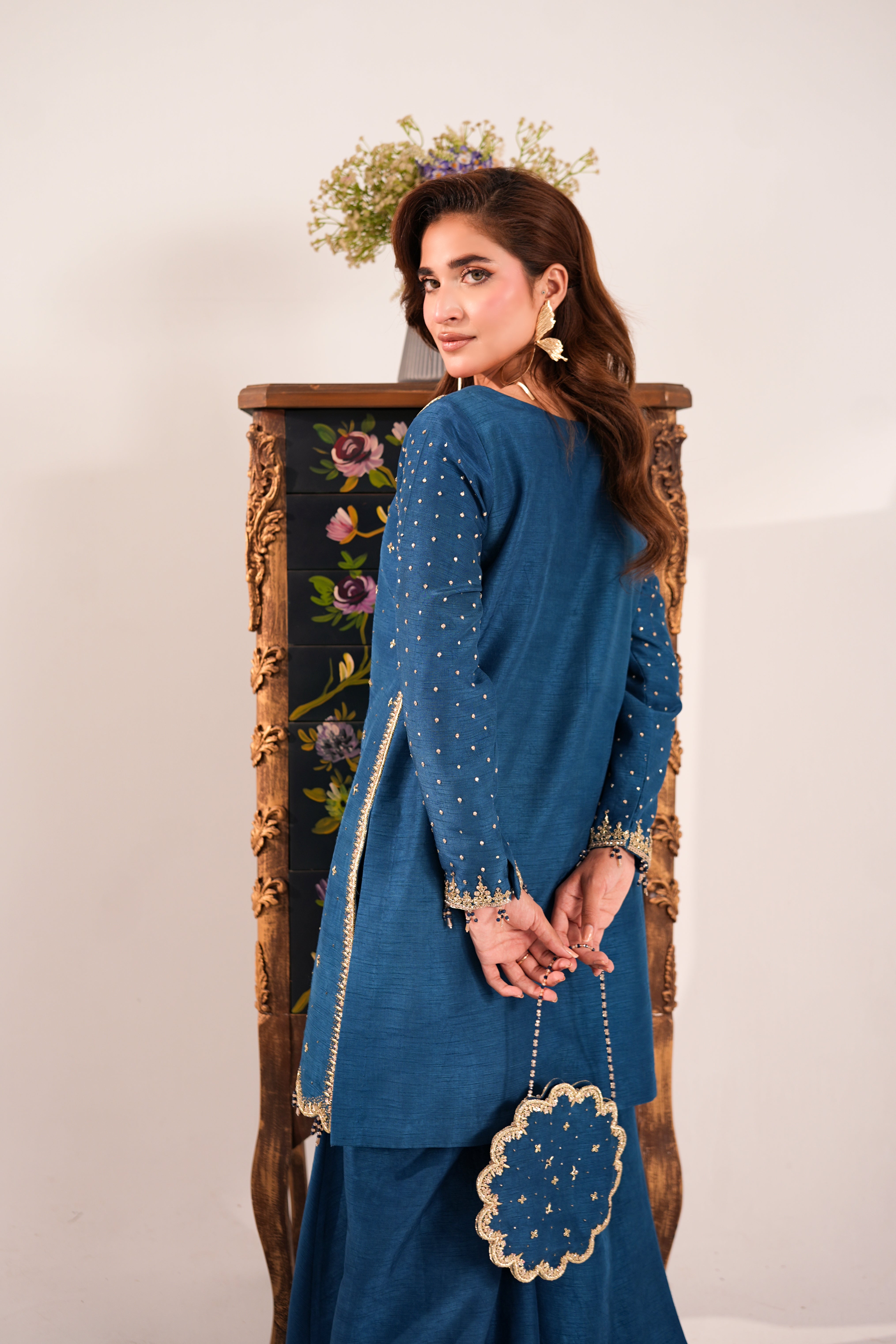 A woman wearing a deep blue traditional outfit with gold embroidery, standing in front of an ornate wooden cabinet against a plain off-white background.