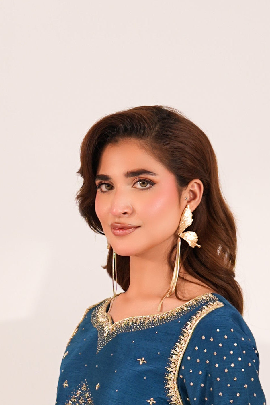 A woman wearing a blue embroidered traditional outfit stands beside a wooden cabinet with floral detailing, with a gray vase of flowers on top, against a plain light background.