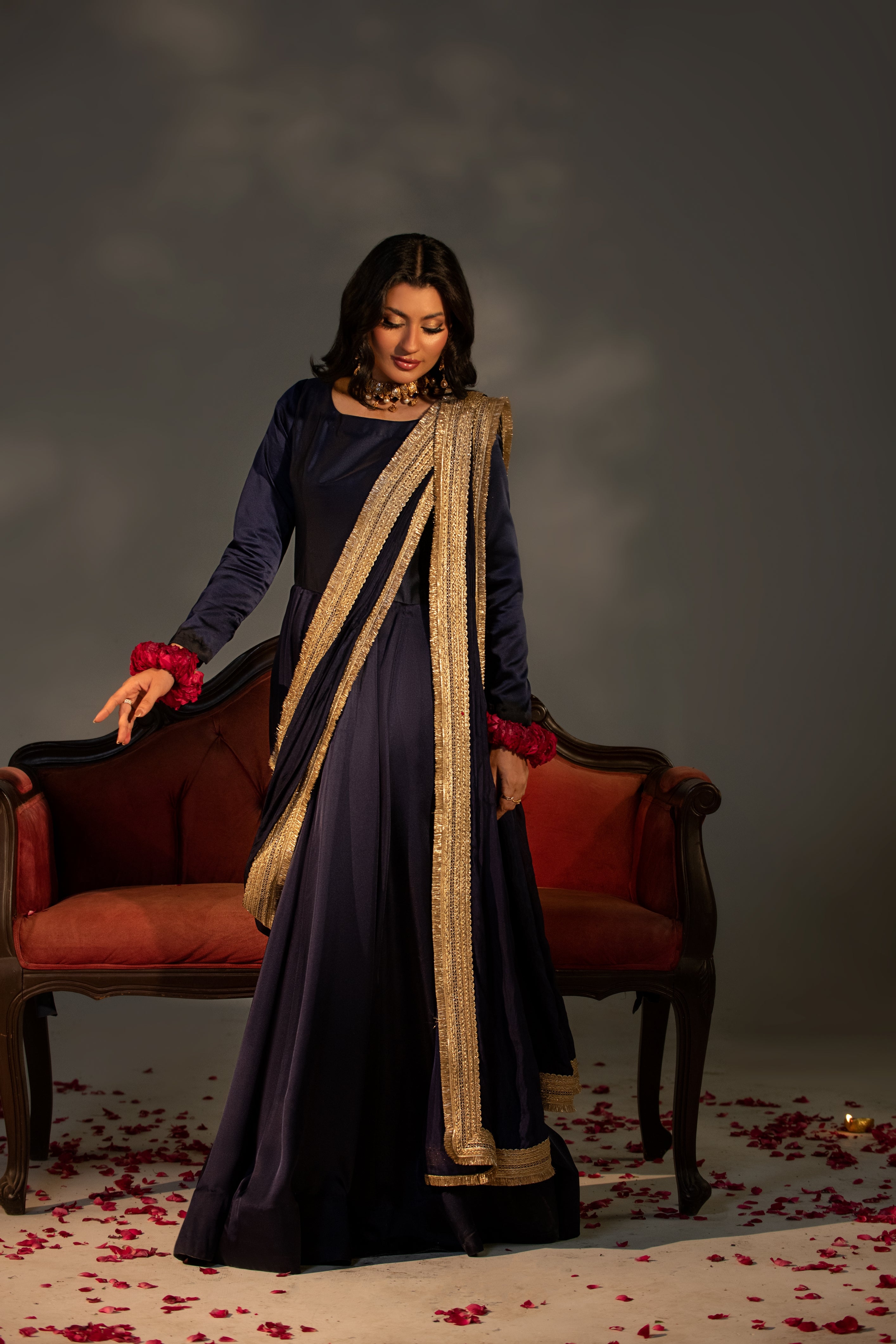 Woman in a dark blue saree with gold border standing in front of a red chair with a gray background.