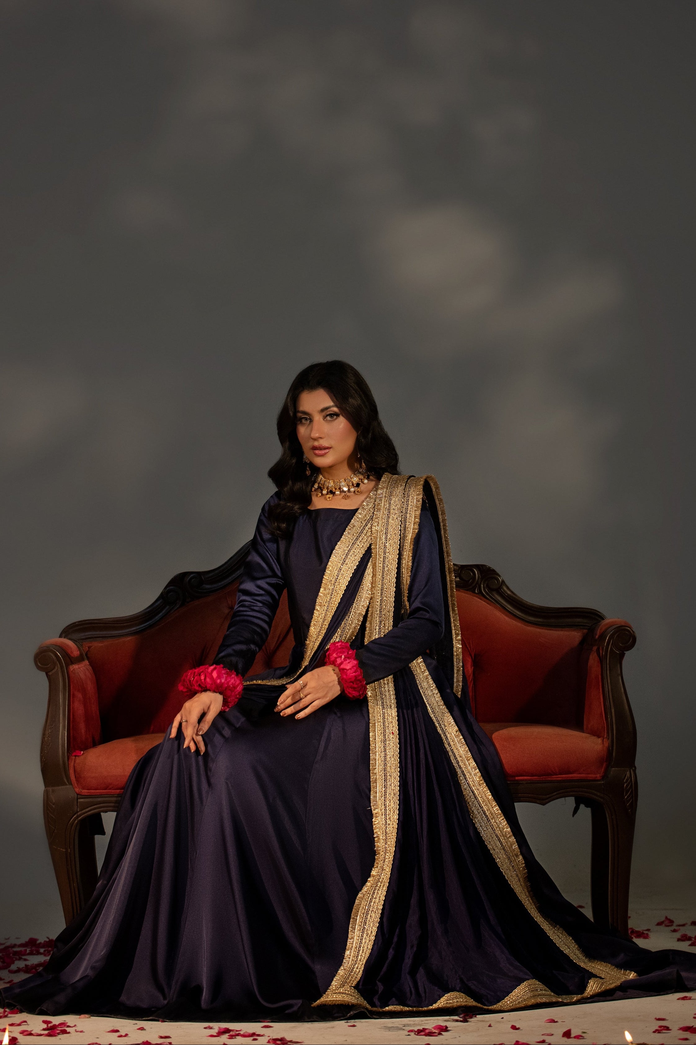 Woman in a dark blue saree with gold border sitting on a red chair against a gray background