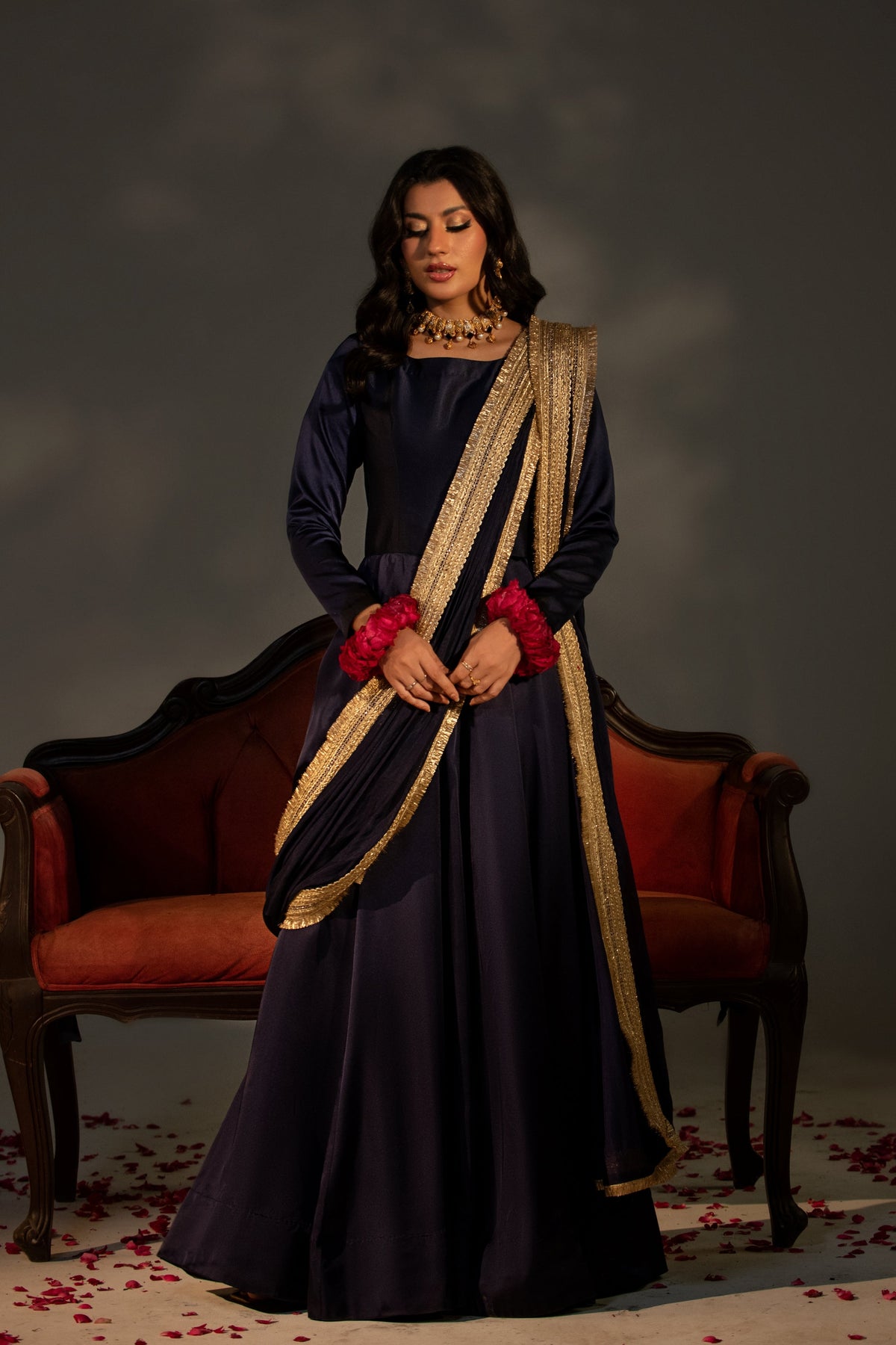 Woman in a dark blue saree with gold border and red flowers, sitting on a red couch against a dark background.