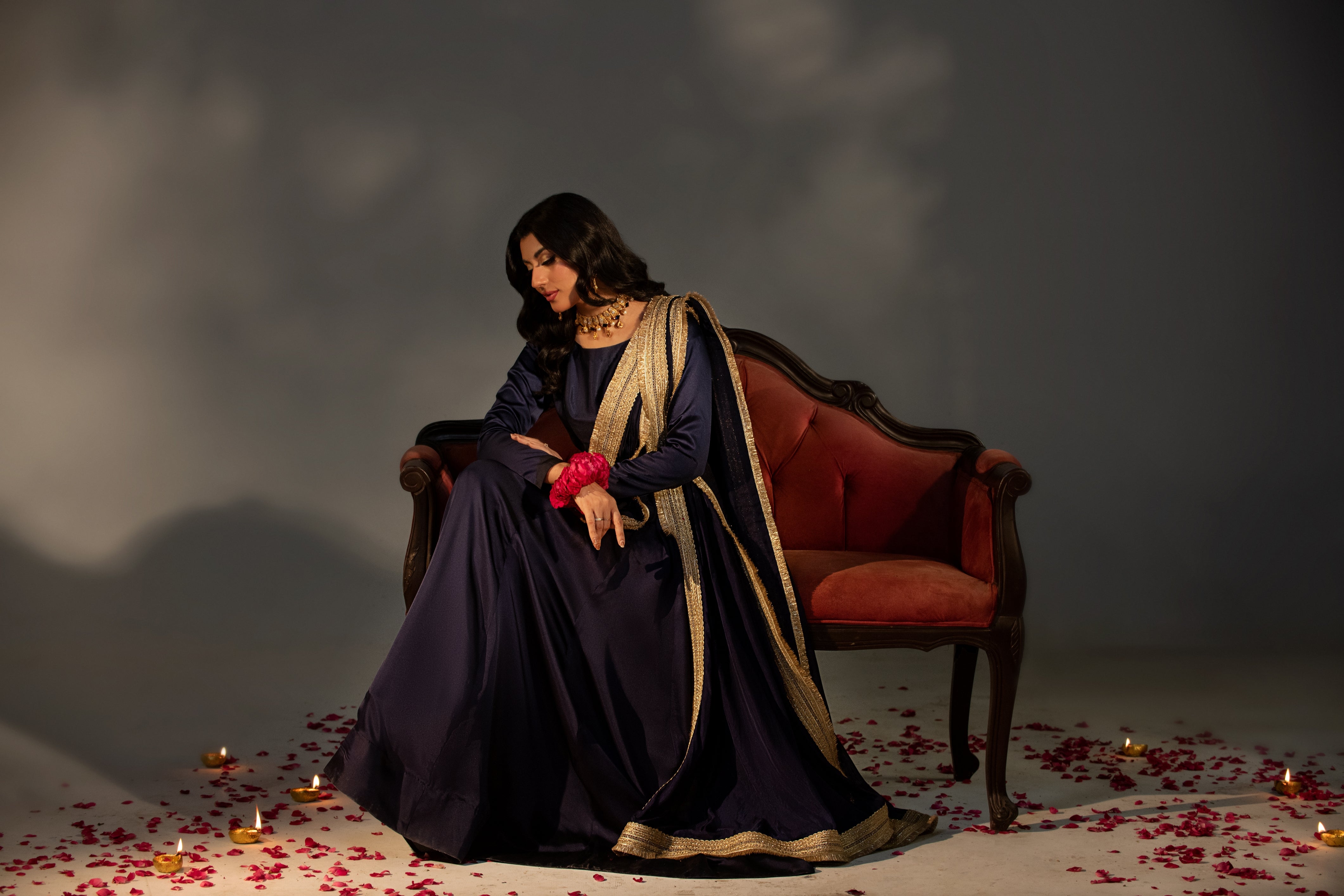 Woman in a dark blue dress with gold accents sitting on a red couch with rose petals on the floor.