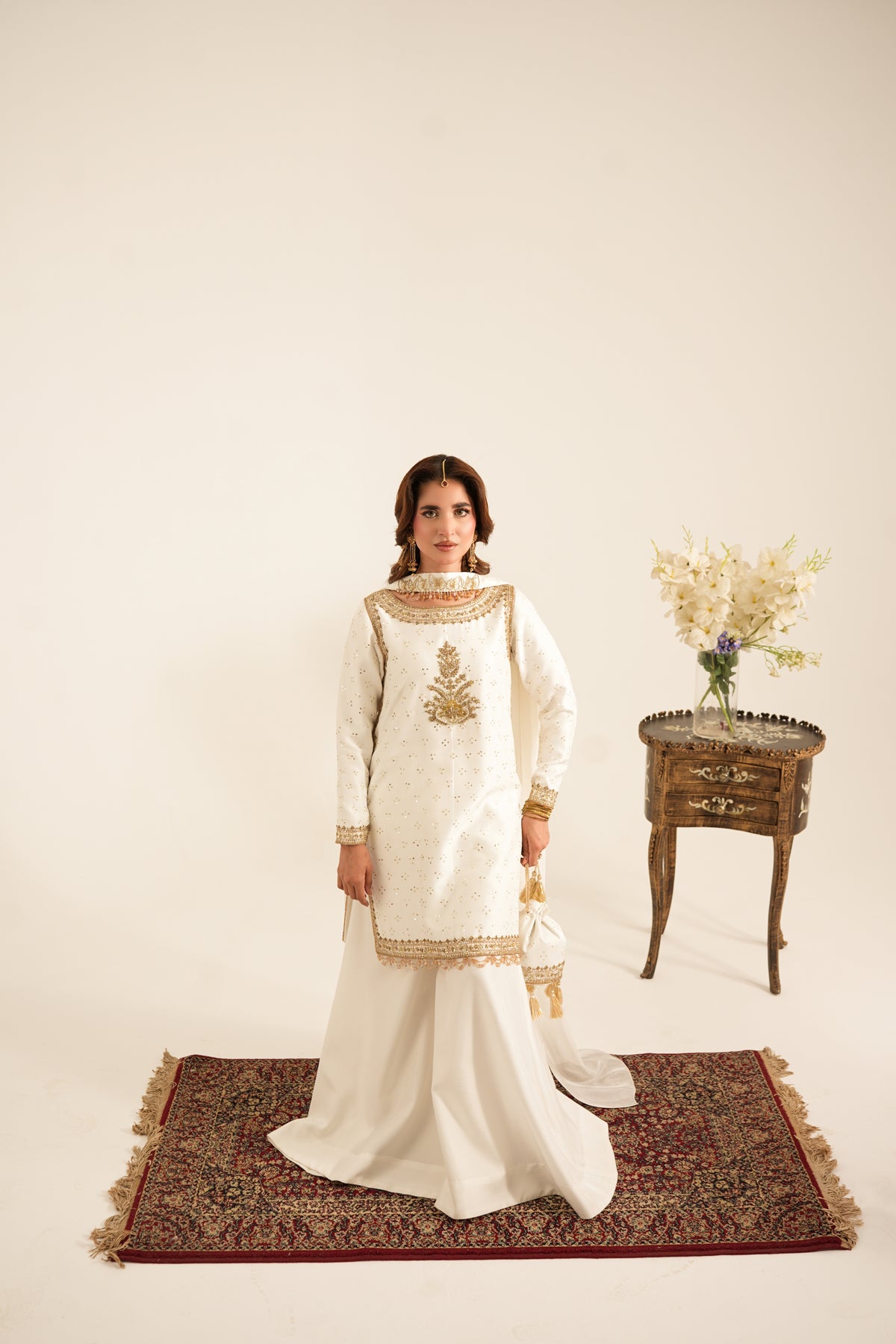 A woman stands on a patterned rug wearing a traditional white embroidered kurta set with a long skirt. She is posed in a studio setting with a small wooden side table holding a vase of flowers in the background.