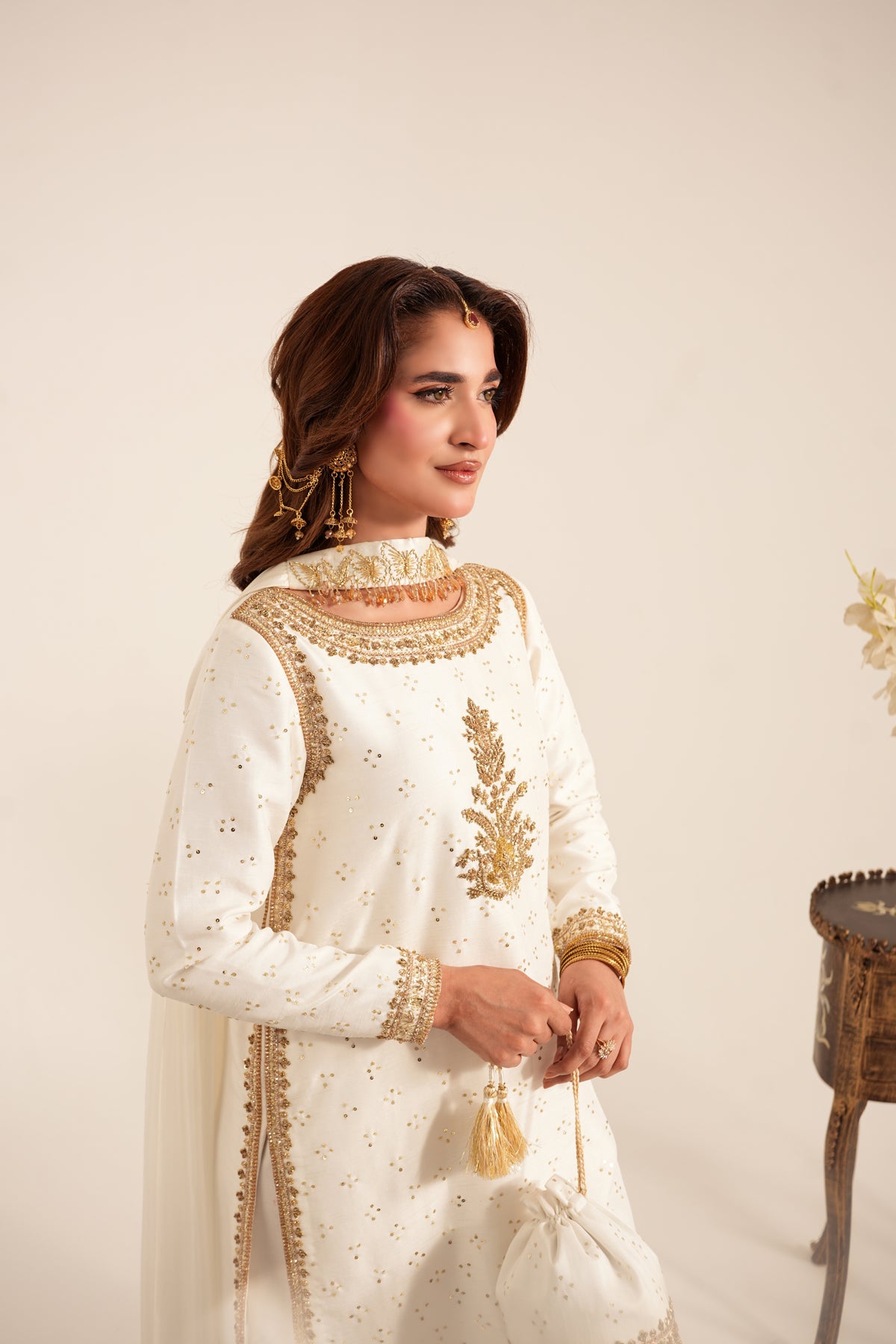 A woman wearing a traditional white embroidered kurta with gold detailing, holding a matching drawstring bag against a warm beige studio background.