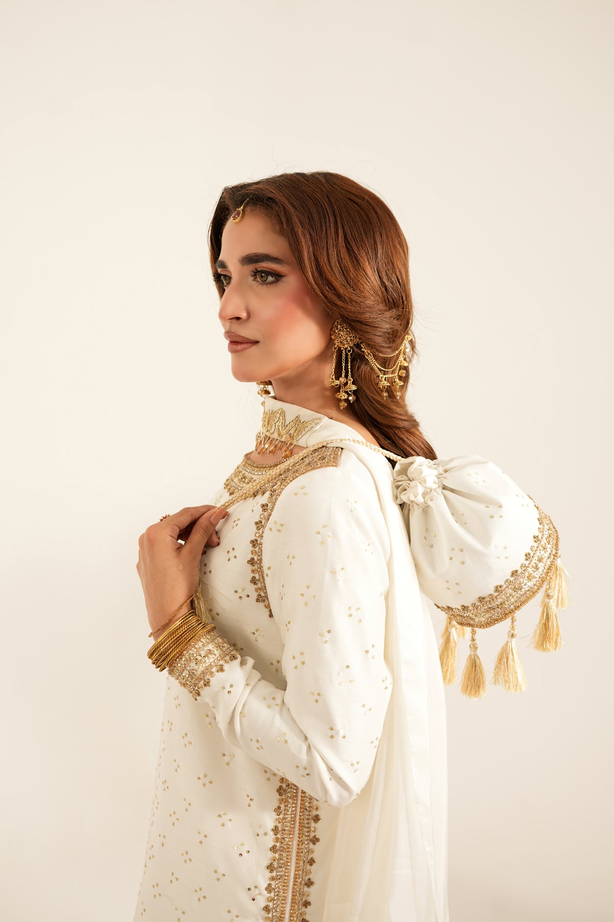 A woman wearing a white traditional outfit with gold embroidery, a matching white dupatta with a large bow and tassels, and gold jewelry, posed against a plain light beige background.