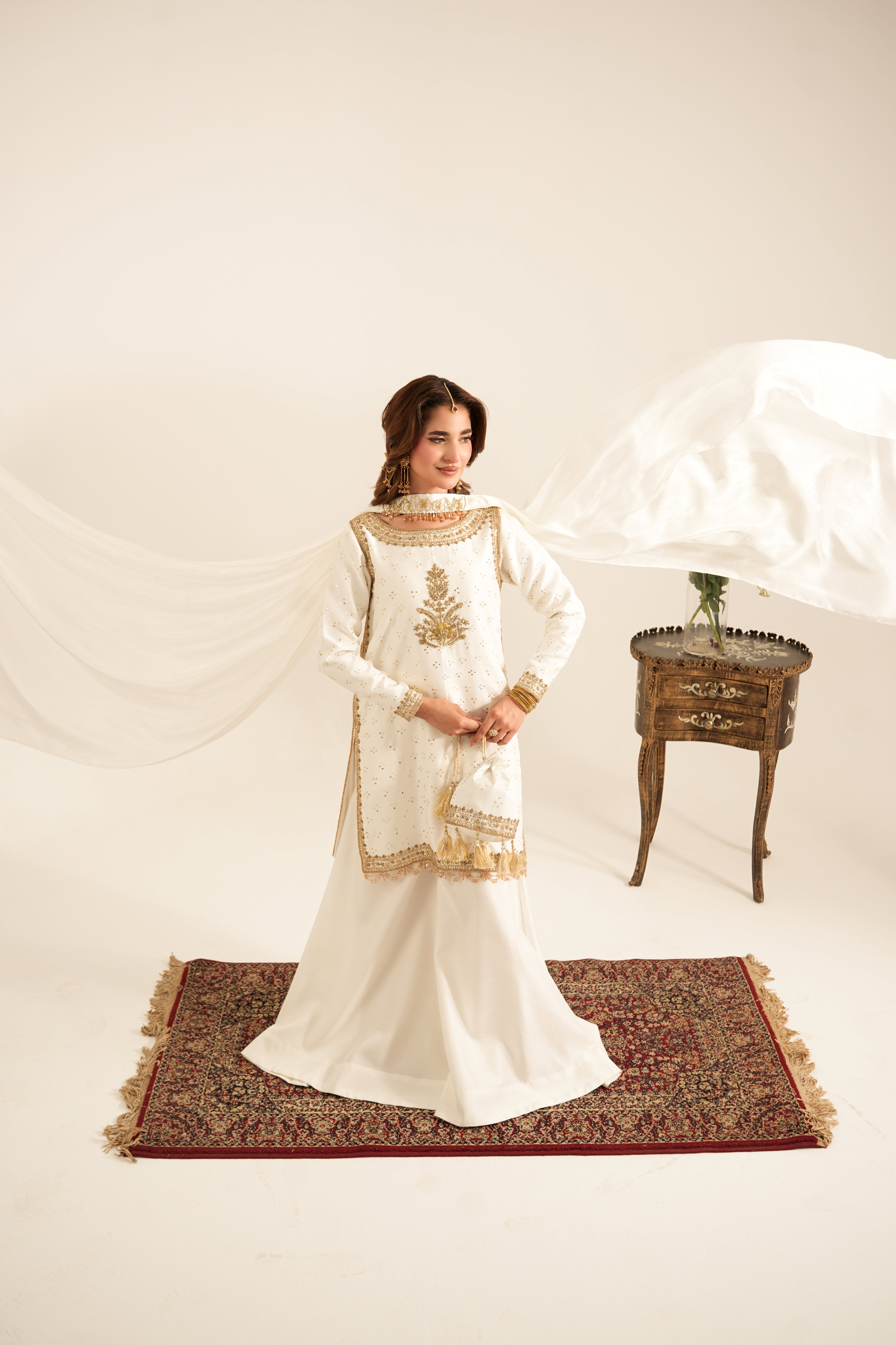 A woman models a traditional white and gold embroidered South Asian outfit, standing on a patterned rug with a draped white fabric and a small wooden side table in the background.