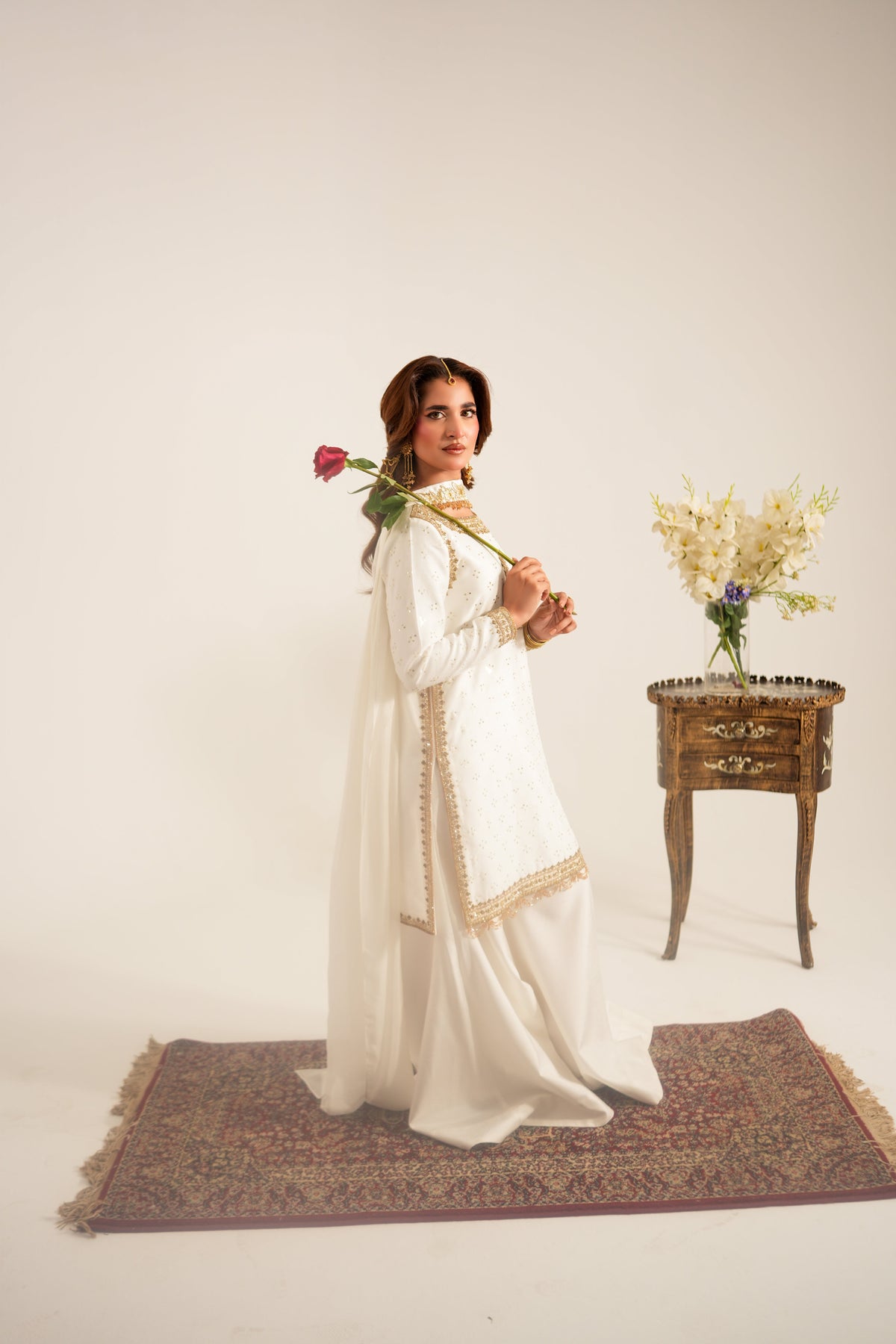 A woman in a white traditional outfit with gold embroidery stands on a patterned rug, holding a red rose. A small wooden side table with yellow flowers is positioned to her right against a plain off-white studio background.