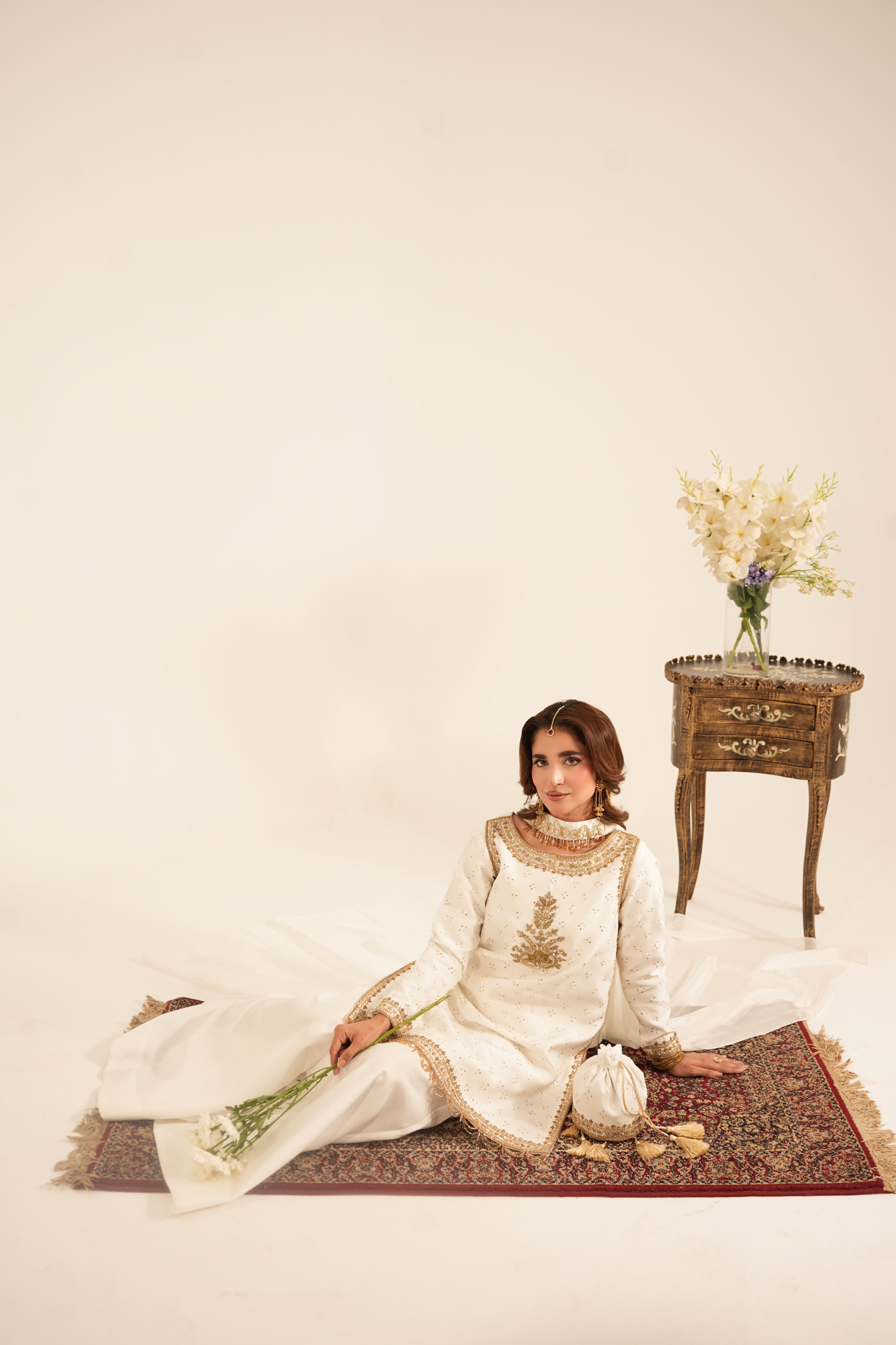 A woman sits cross-legged on a patterned rug in a studio setting, wearing a long white traditional outfit with gold embroidery. A small wooden side table with a vase of flowers stands to her right against a plain cream backdrop.
