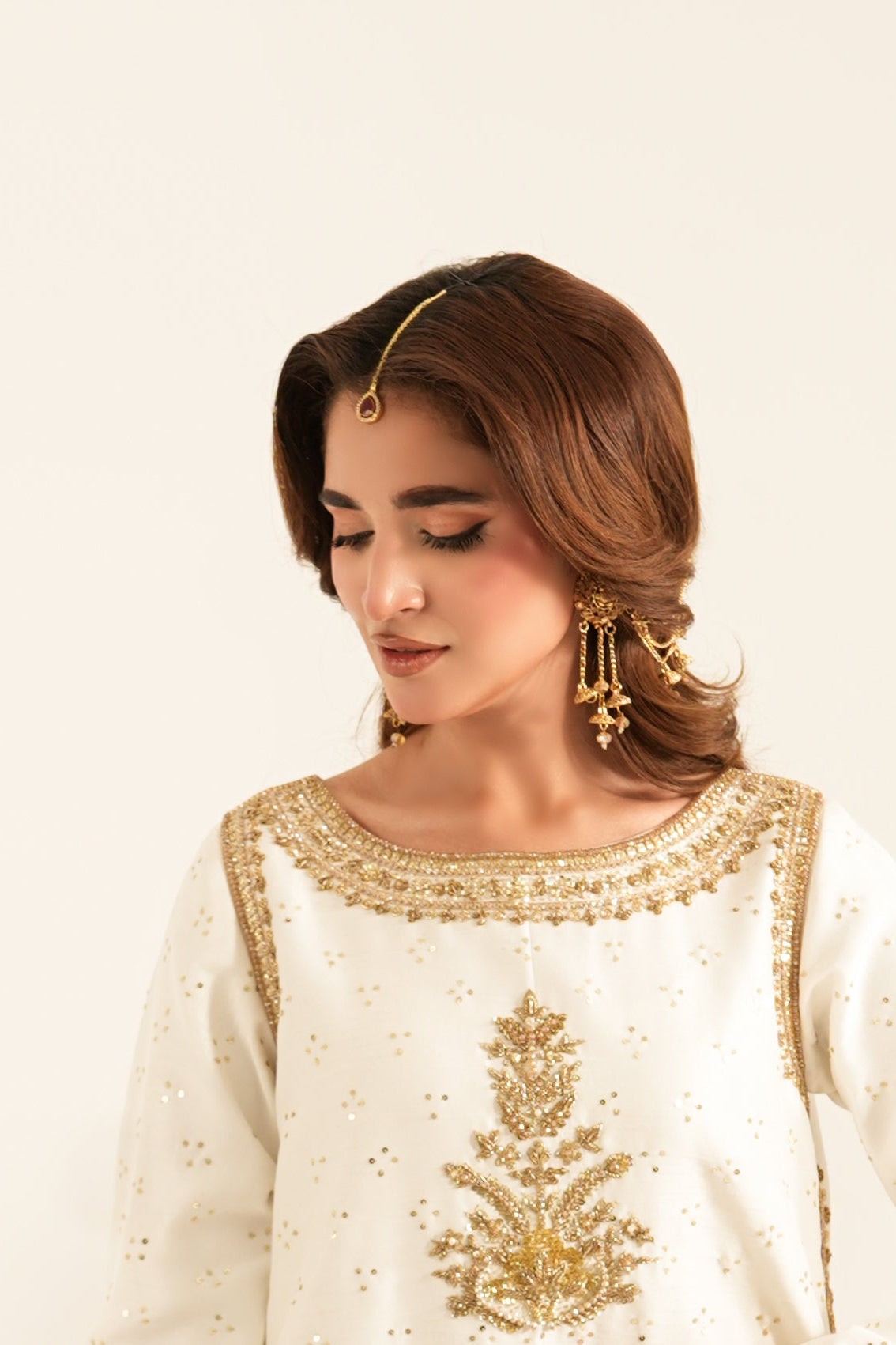 A woman wearing a white embroidered traditional outfit with gold detailing, standing against a plain light beige background next to a small vase of white flowers.