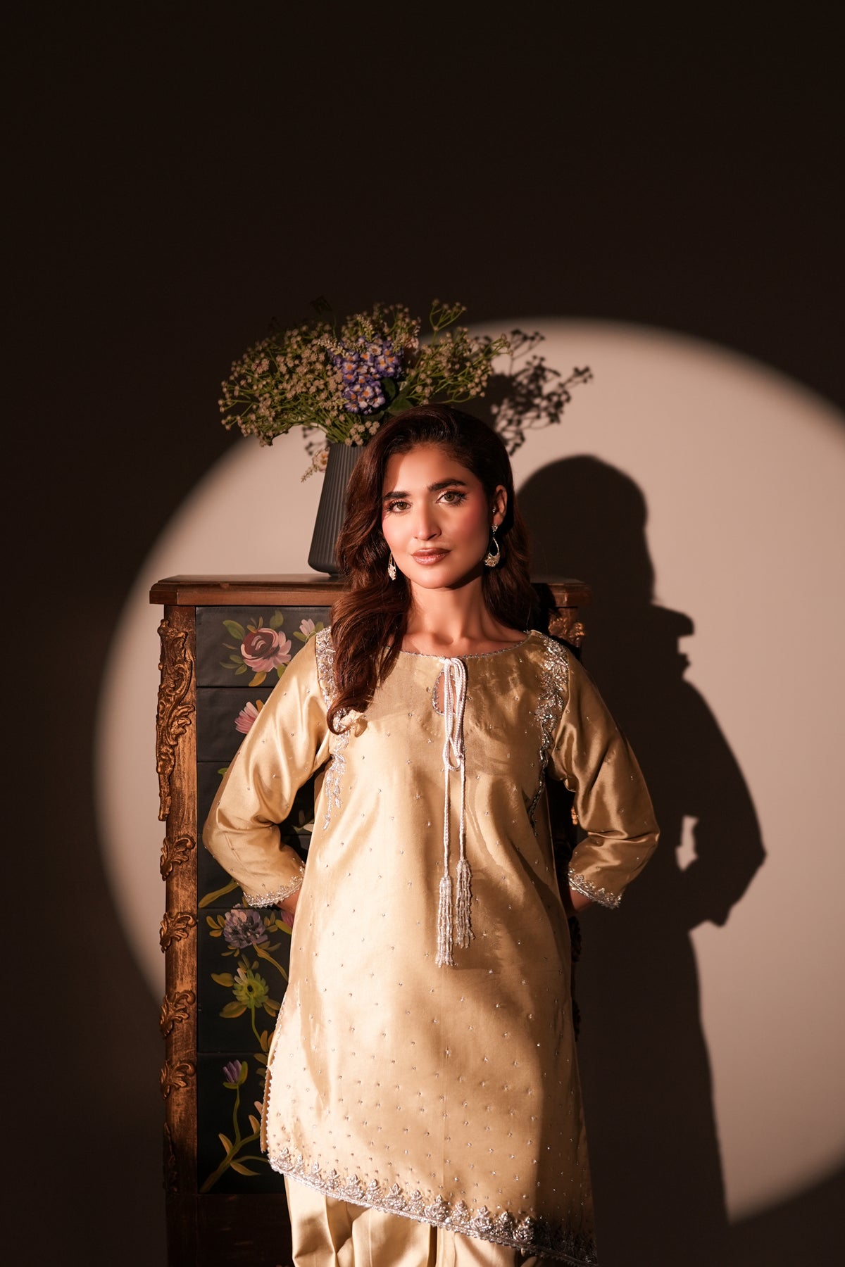 A woman models a traditional beige embroidered kurta set with matching trousers, standing in front of a dark wall with dramatic spotlighting and a floral arrangement in the background.