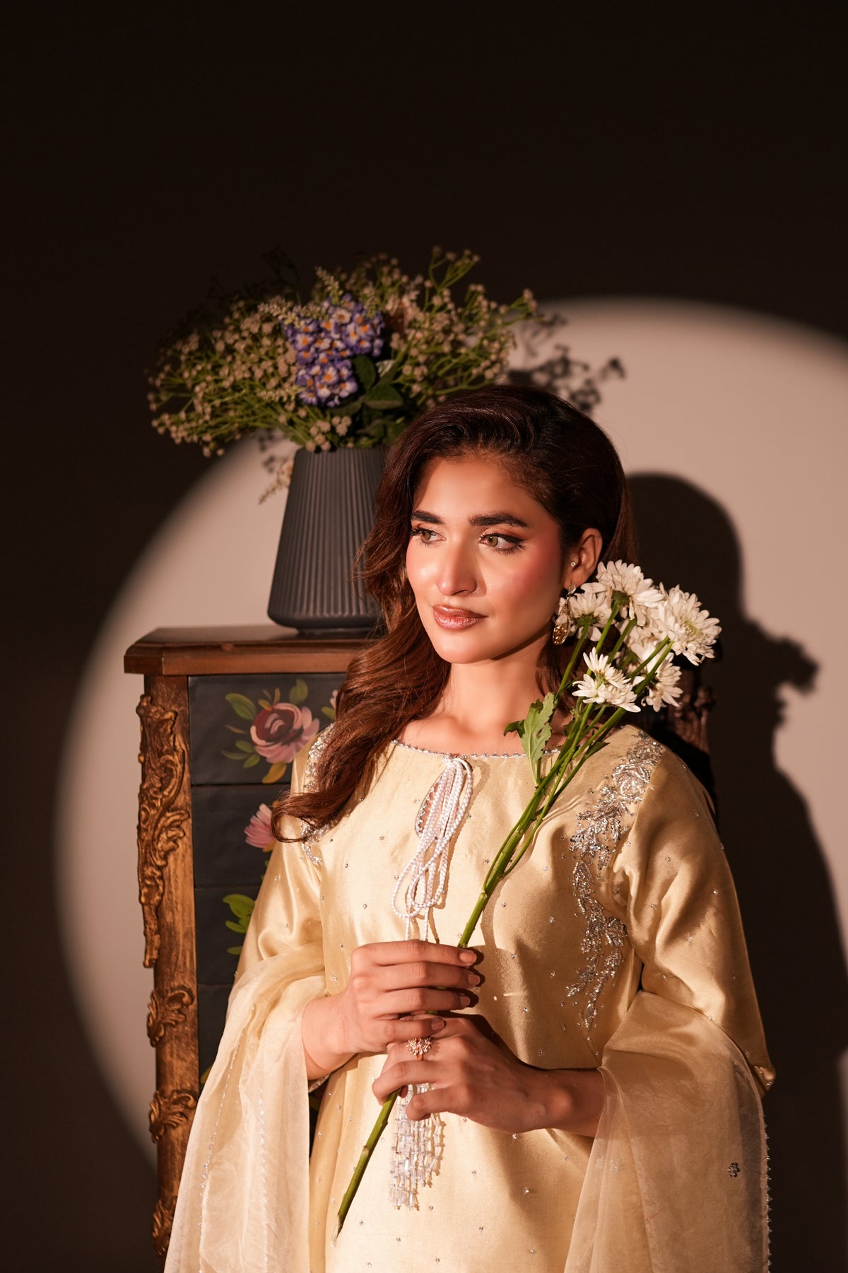 A woman wearing a cream-colored embroidered traditional outfit holds white flowers, standing in front of a dark background with a spotlight effect and a decorative cabinet.
