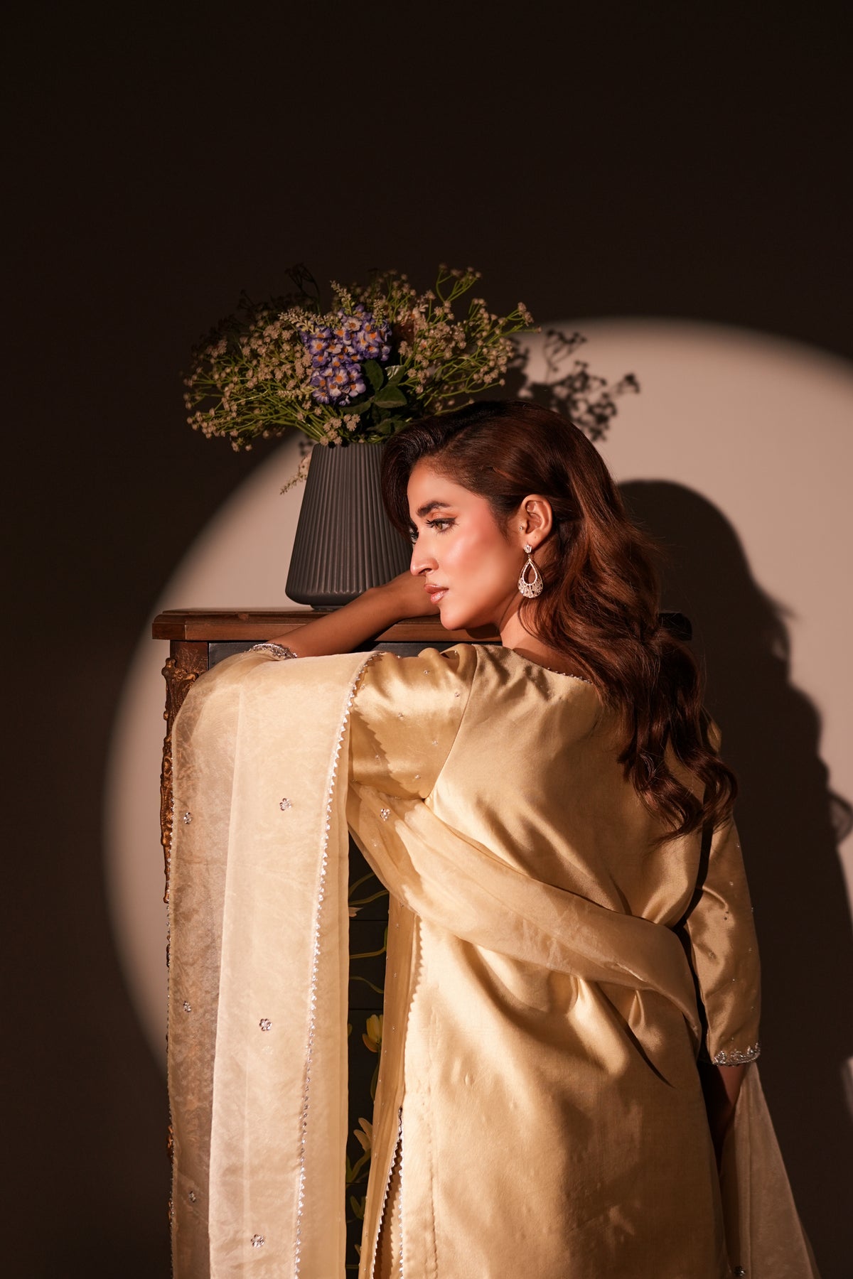 A woman in a shimmering gold traditional outfit with a matching sheer dupatta, posed in a dramatic studio setting with a spotlight and a vase of flowers in the background.