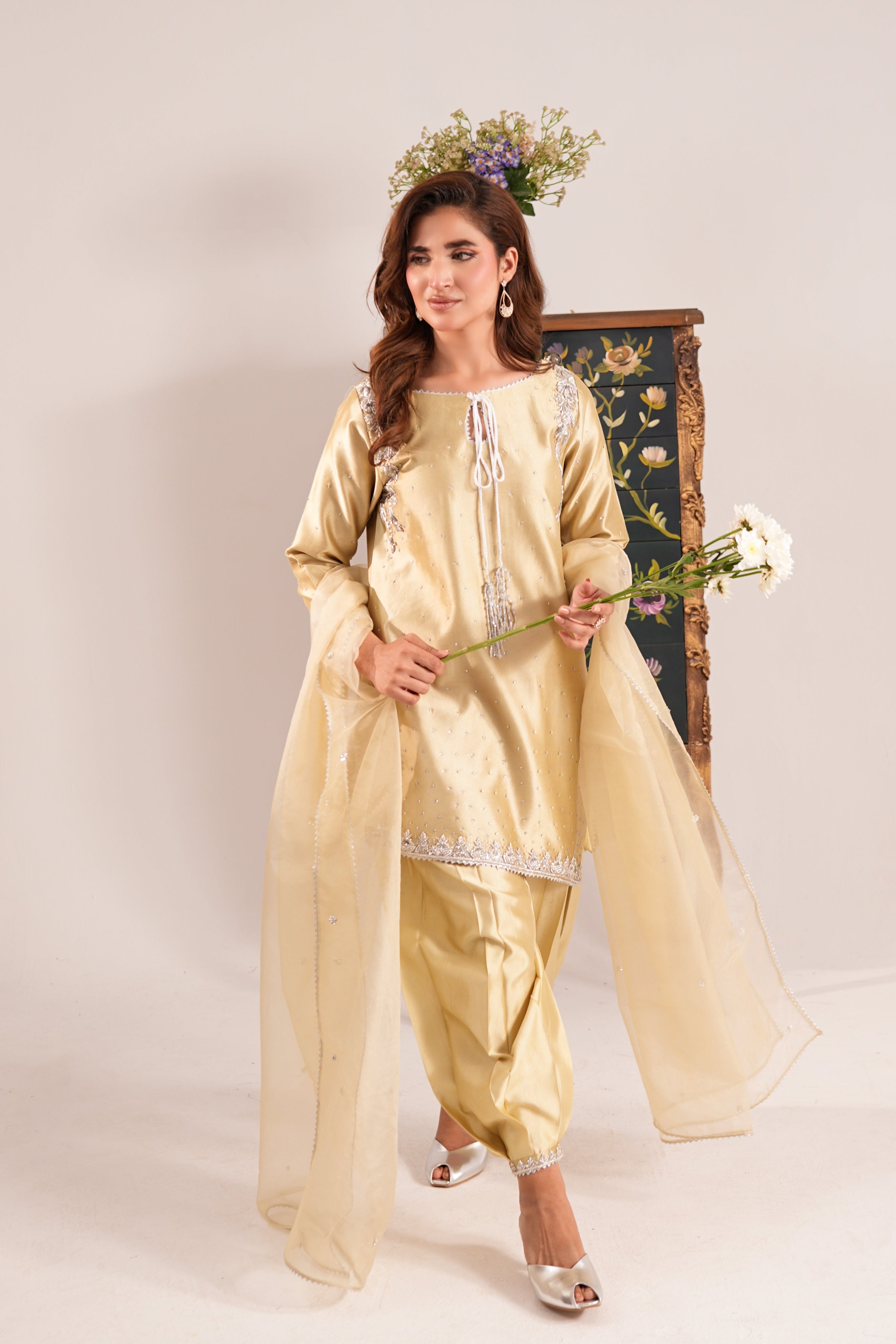 A woman models a traditional South Asian outfit consisting of a light gold kurta, matching pants, and a sheer dupatta, styled with a floral headpiece and holding white flowers against a neutral studio backdrop.