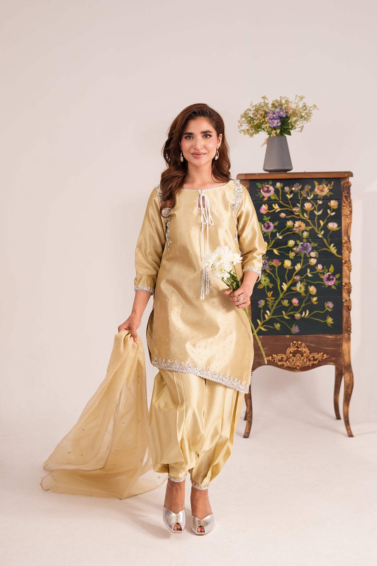 A woman models a traditional Indian salwar kameez suit in a light gold/champagne color with silver embroidery. She is standing in a studio setting next to an ornate dark green floral cabinet, holding a matching sheer dupatta and a small bouquet of white flowers.