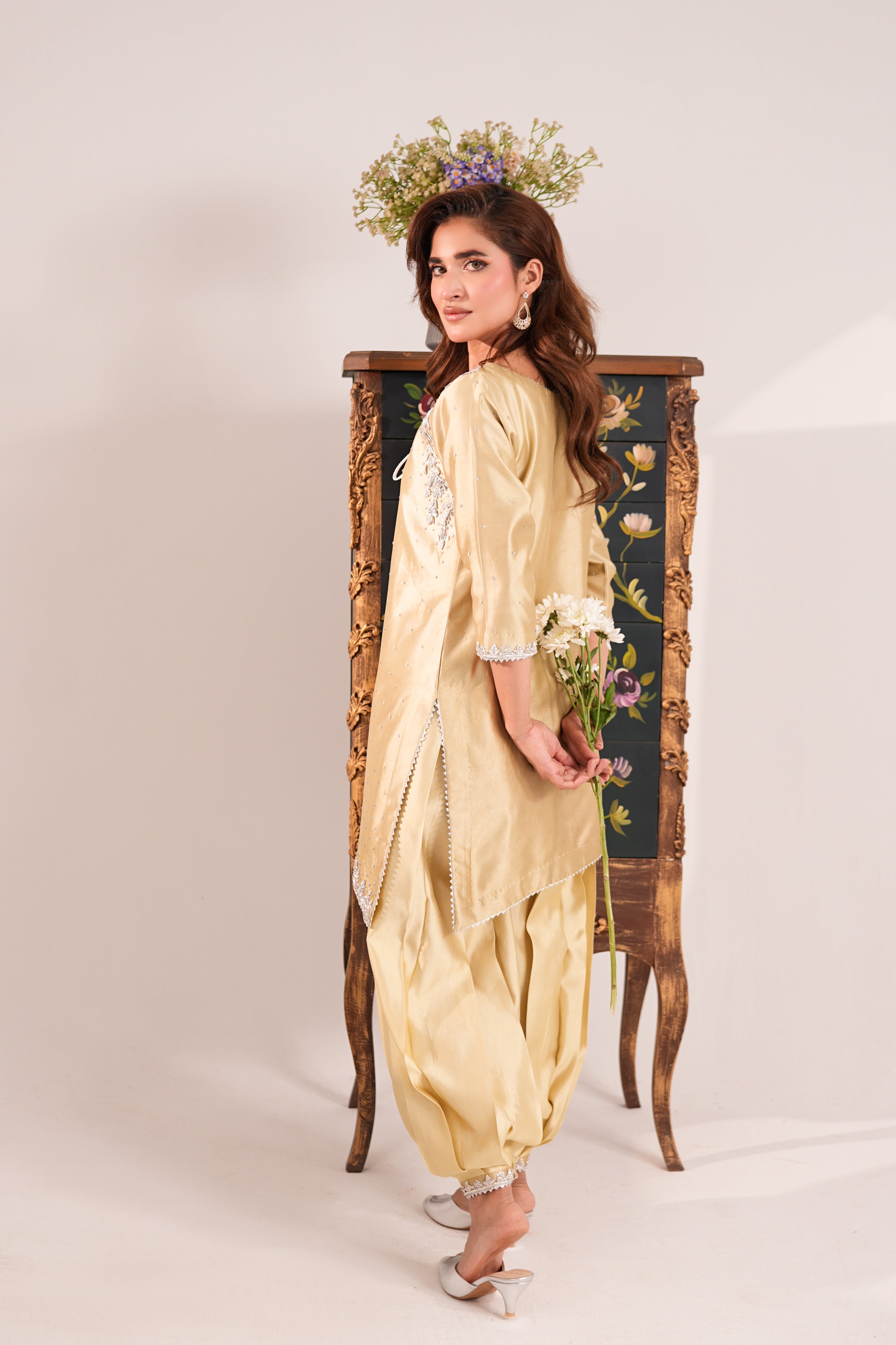 A woman models a traditional light yellow salwar suit set consisting of a kurta and matching pants, standing in front of an ornate wooden mirror. She is wearing a floral headpiece and holding a small bouquet of white flowers.